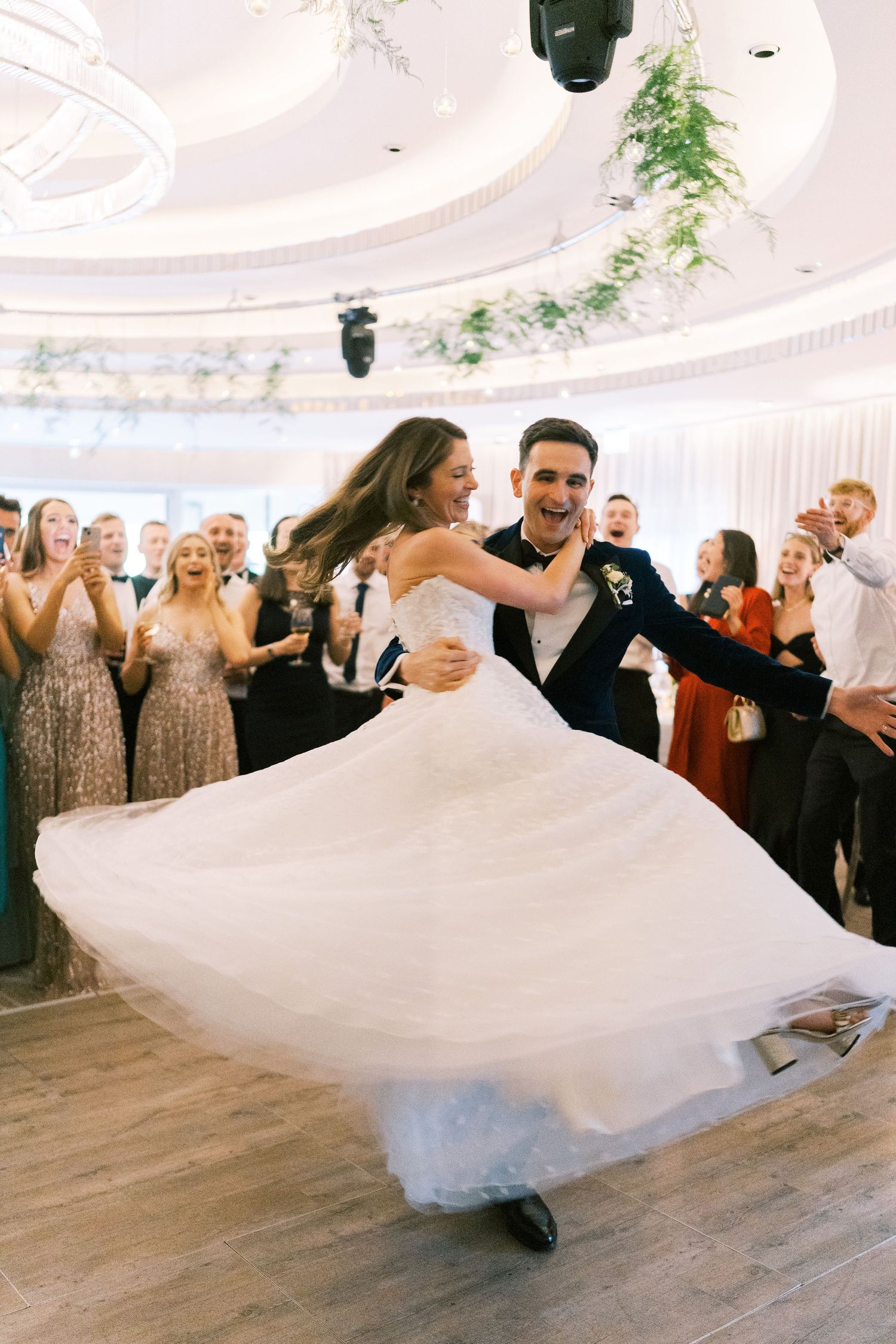 Bride and groom share joyful first dance, wedding dress swirling, guests cheering in elegant ballroom.