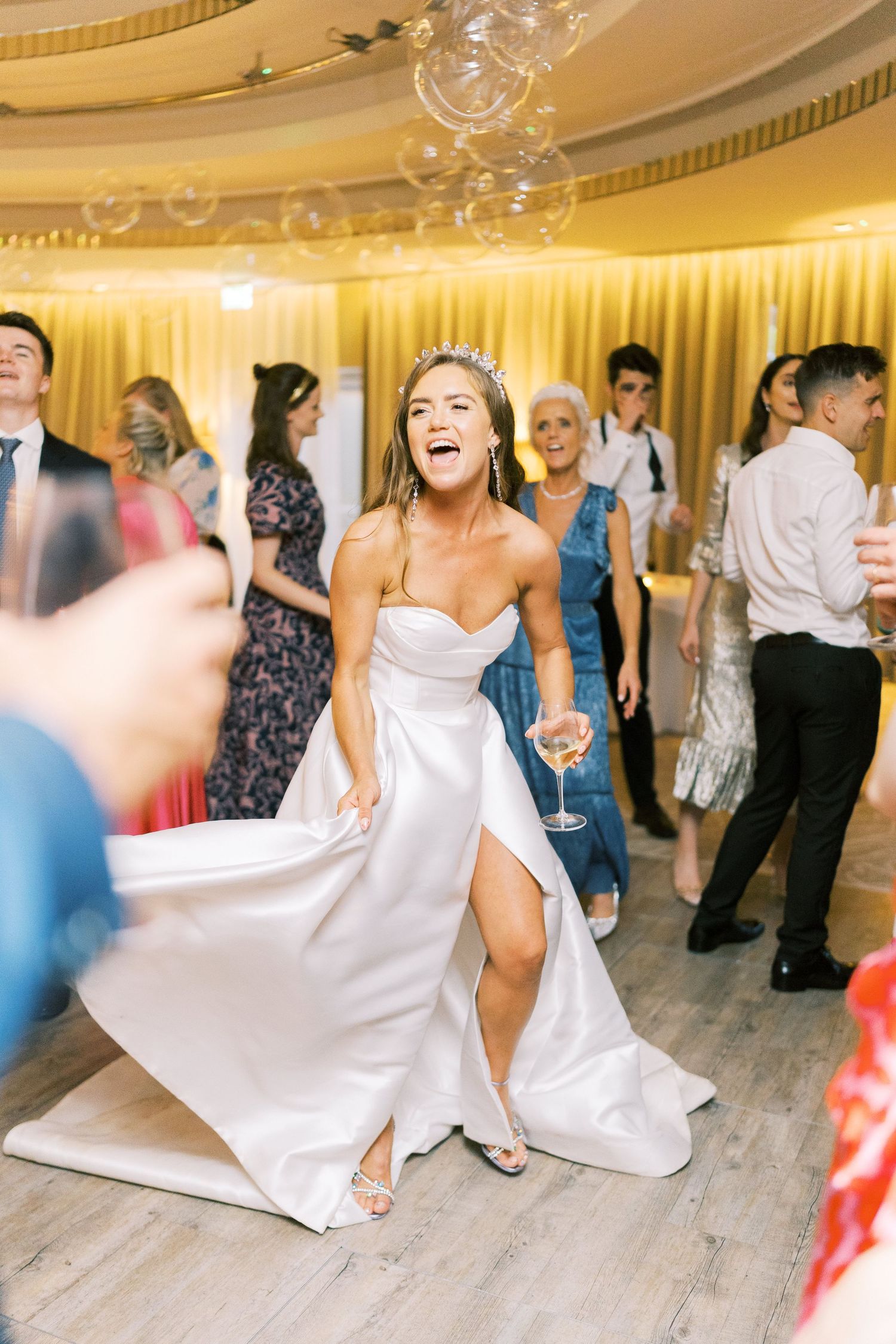 Radiant bride in strapless white gown dances with champagne glass at lively wedding reception.