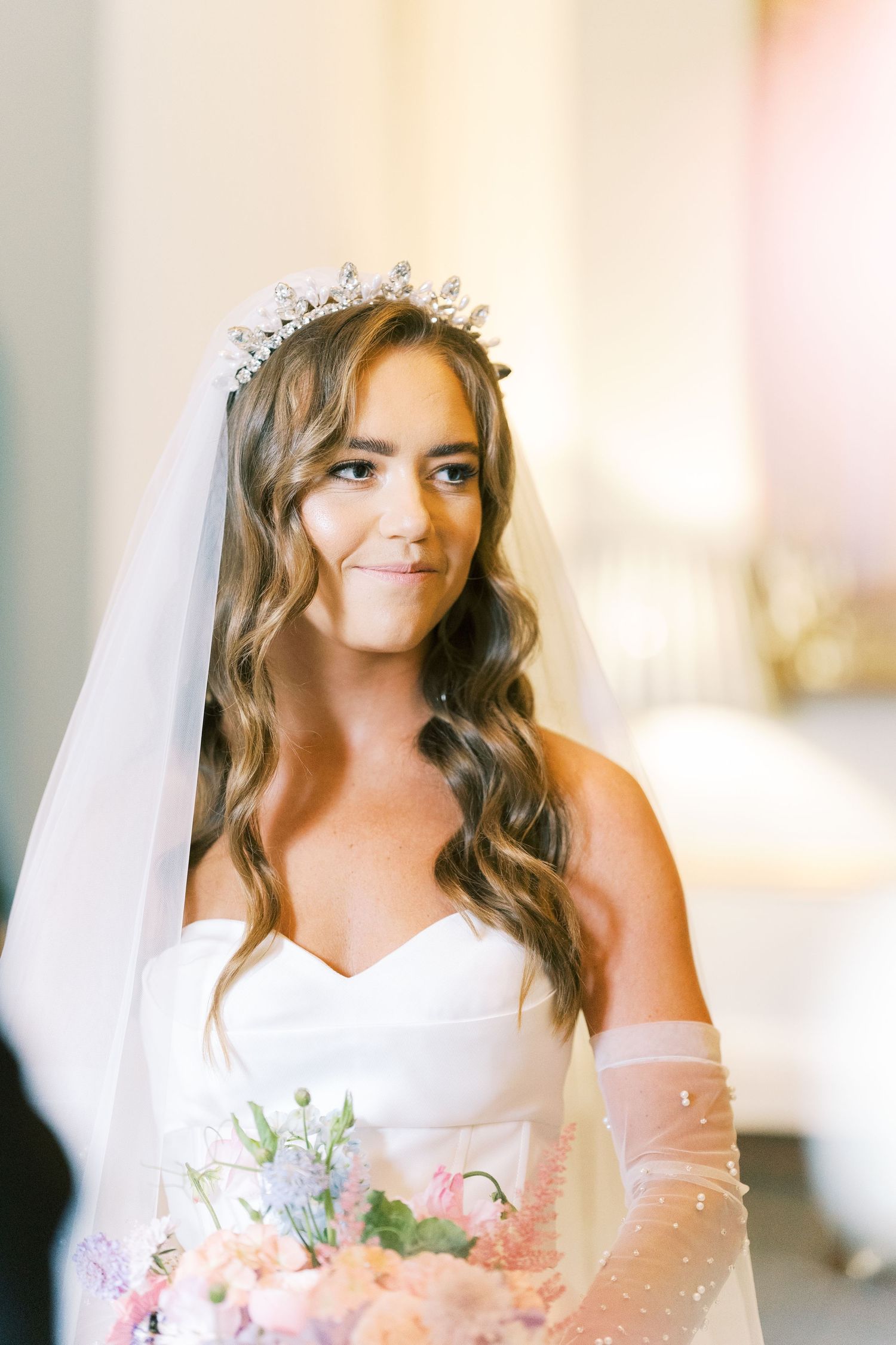 Bride in strapless white gown, crystal tiara, veil, and pink floral bouquet smiles softly indoors.