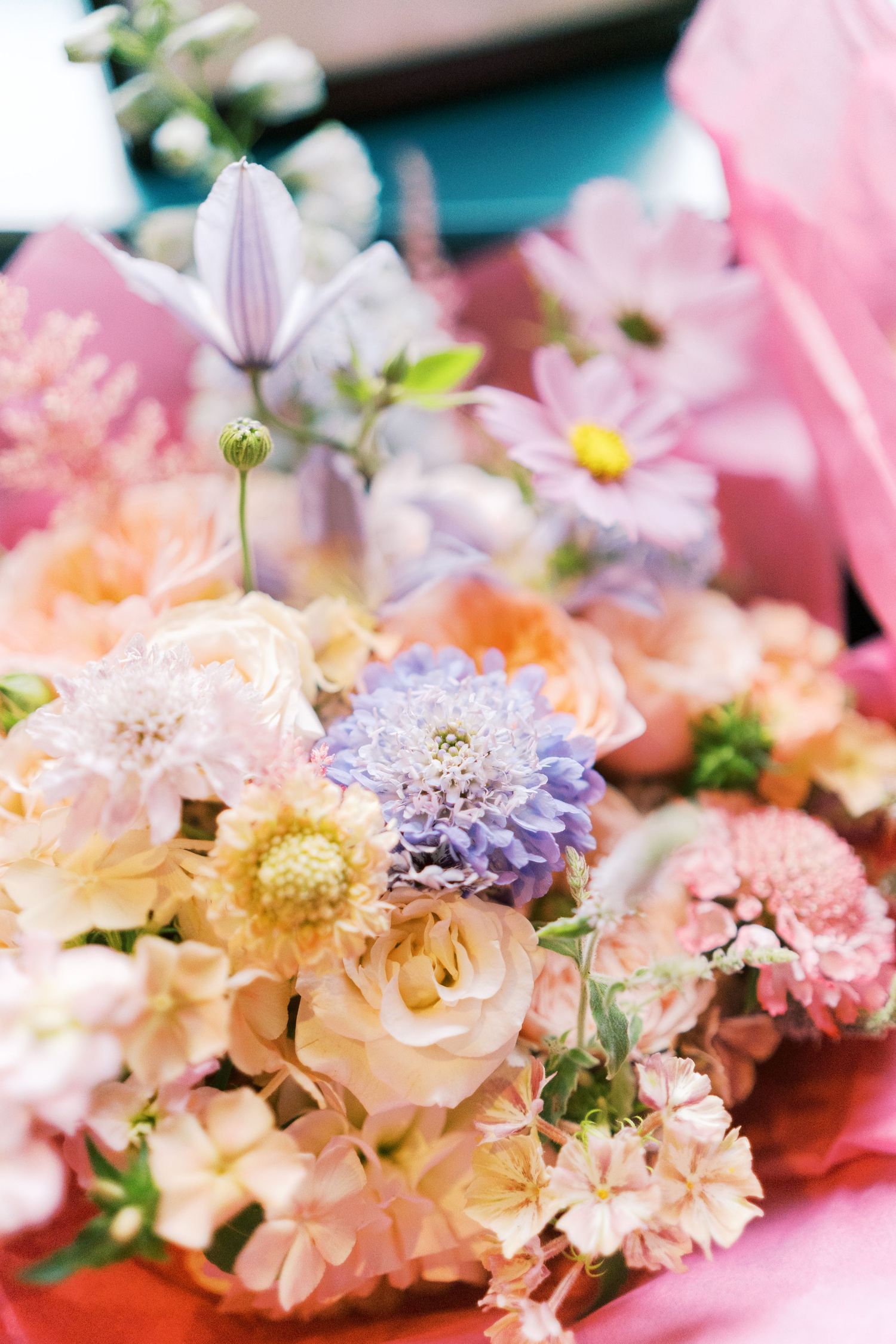Pastel wedding bouquet featuring roses, dahlias, and cosmos in soft pink, peach, lavender, and white tones wrapped in pink paper.