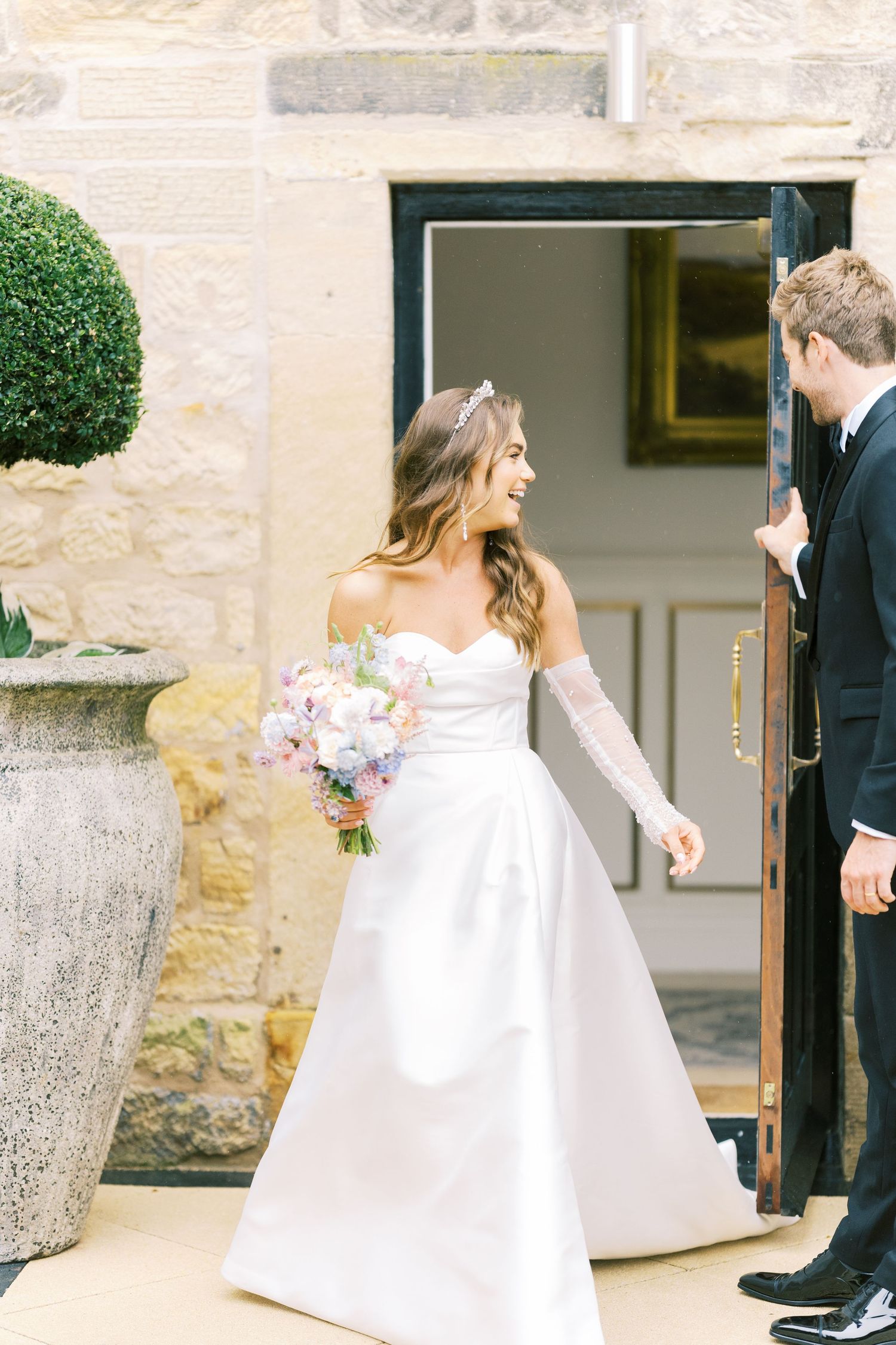 Smiling bride in white off-shoulder gown with sheer gloves holds pastel bouquet at elegant stone venue entrance.