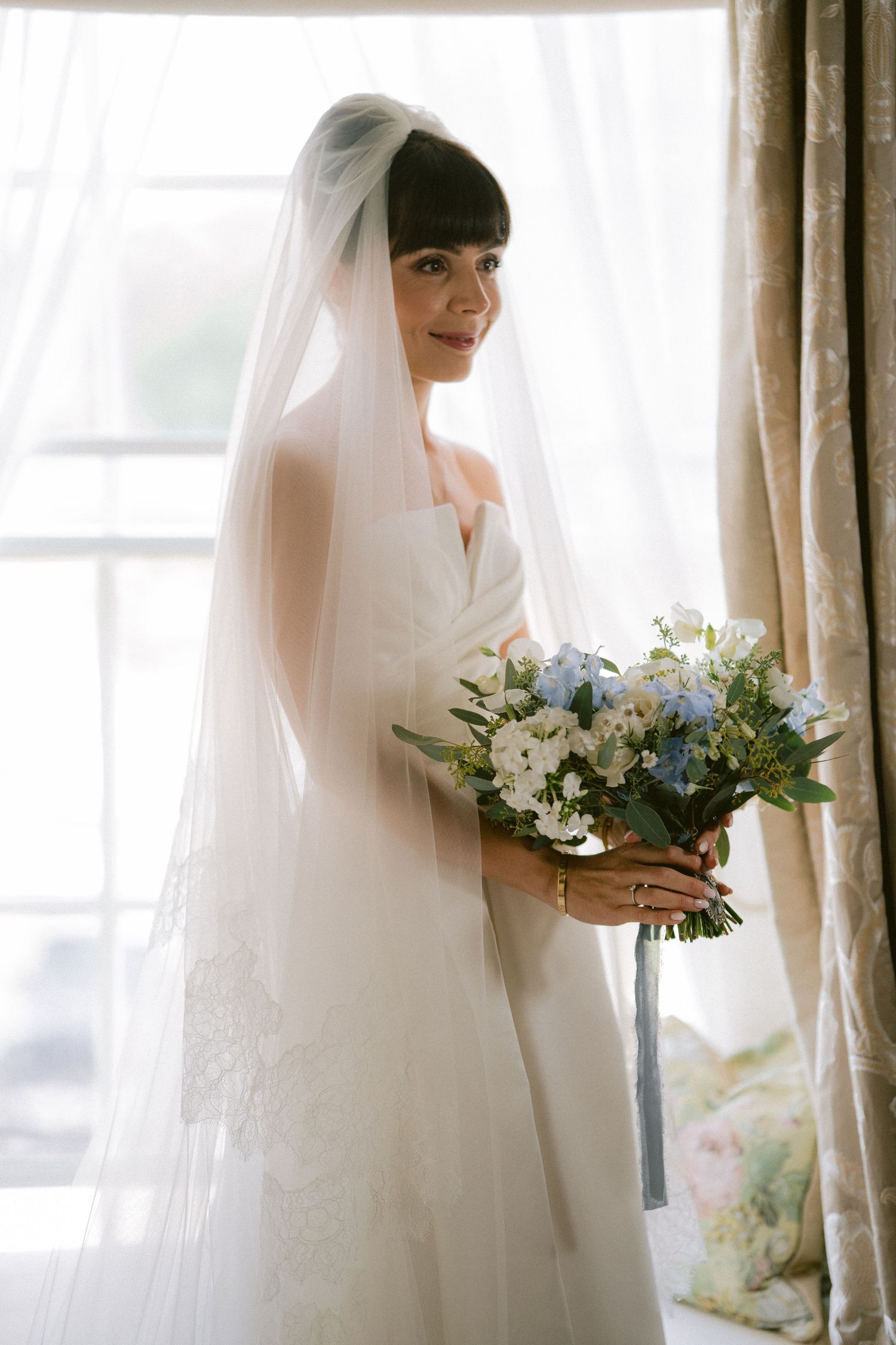 Bride in white lace gown and veil holding blue and white floral bouquet standing by window.