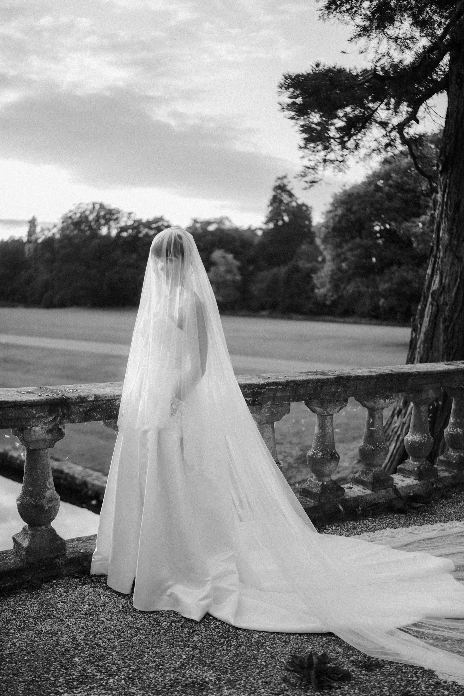 Black and white portrait of bride in long veil standing at stone balustrade in garden.