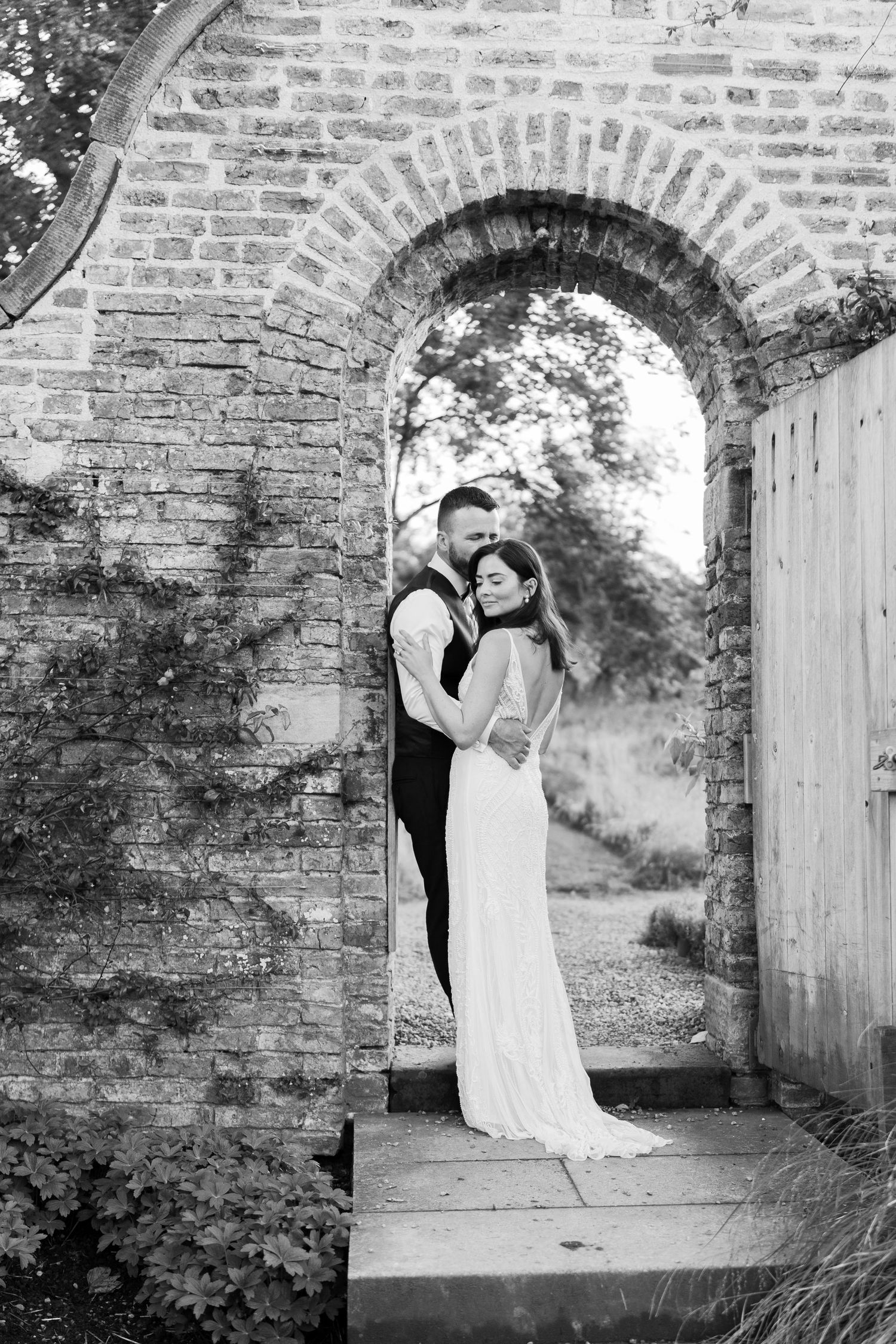 Black and white photo of a bride and groom embracing under a rustic brick archway at their wedding.