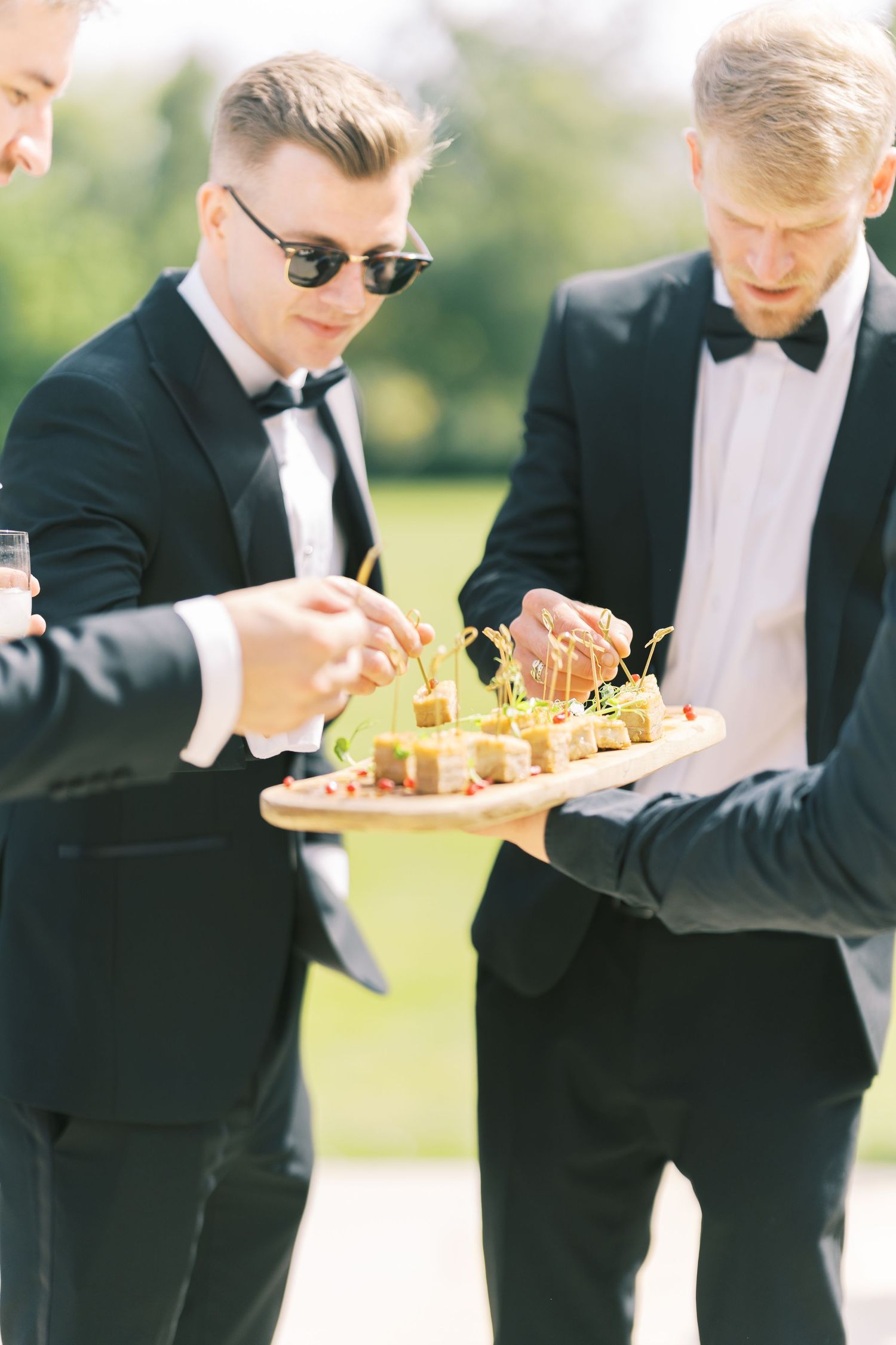 Men in black tuxedos enjoying elegant appetizers on a wooden tray at an outdoor formal event.