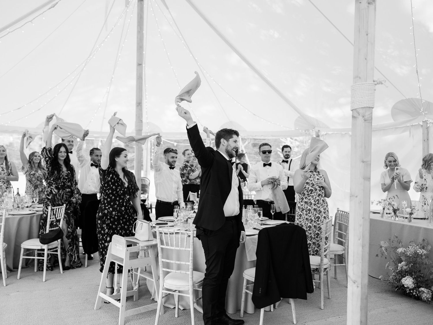 Black and white wedding reception photo of guests celebrating under a marquee tent, raising hands and applauding.