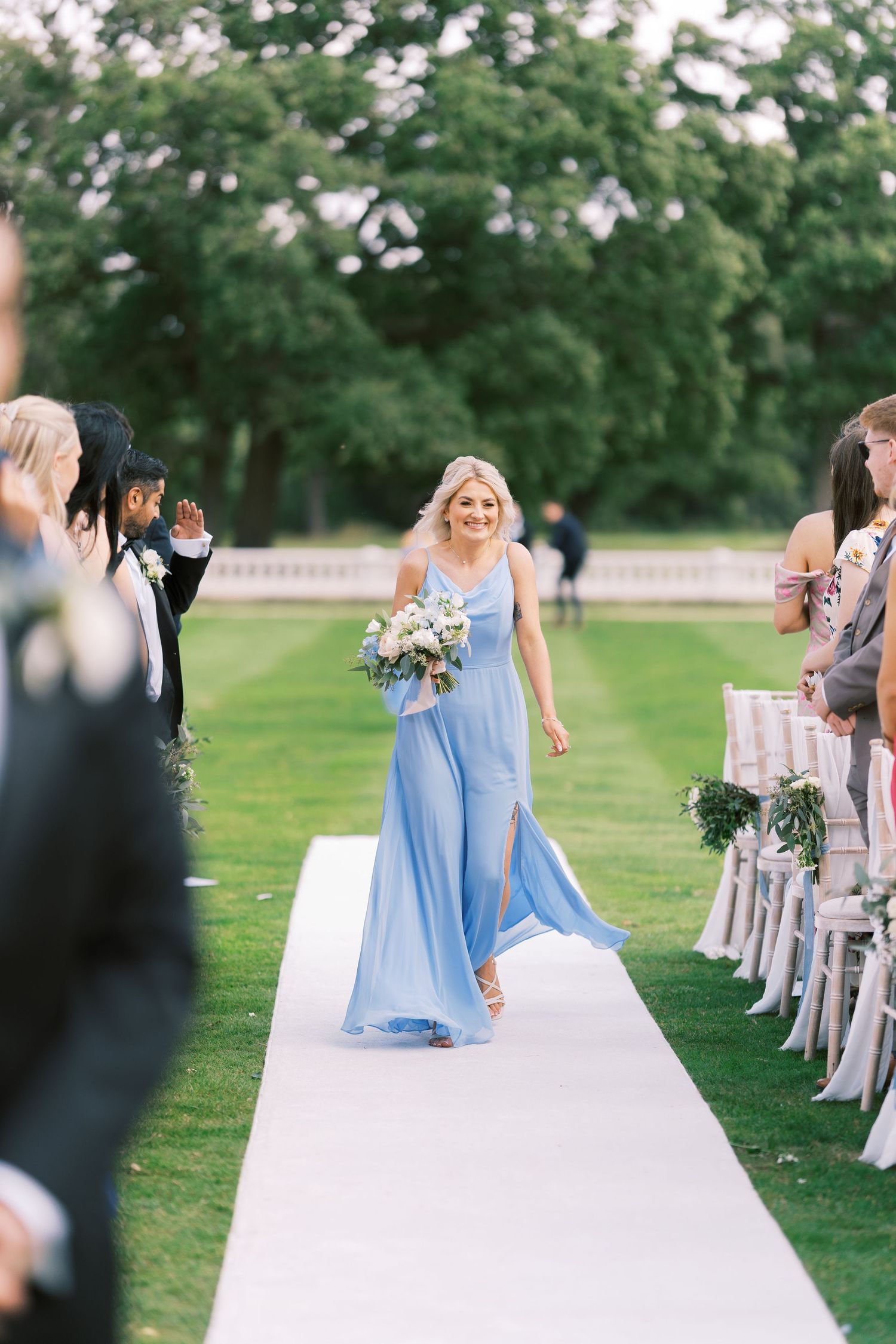 Smiling bridesmaid in light blue gown walks down outdoor wedding aisle carrying white floral bouquet.