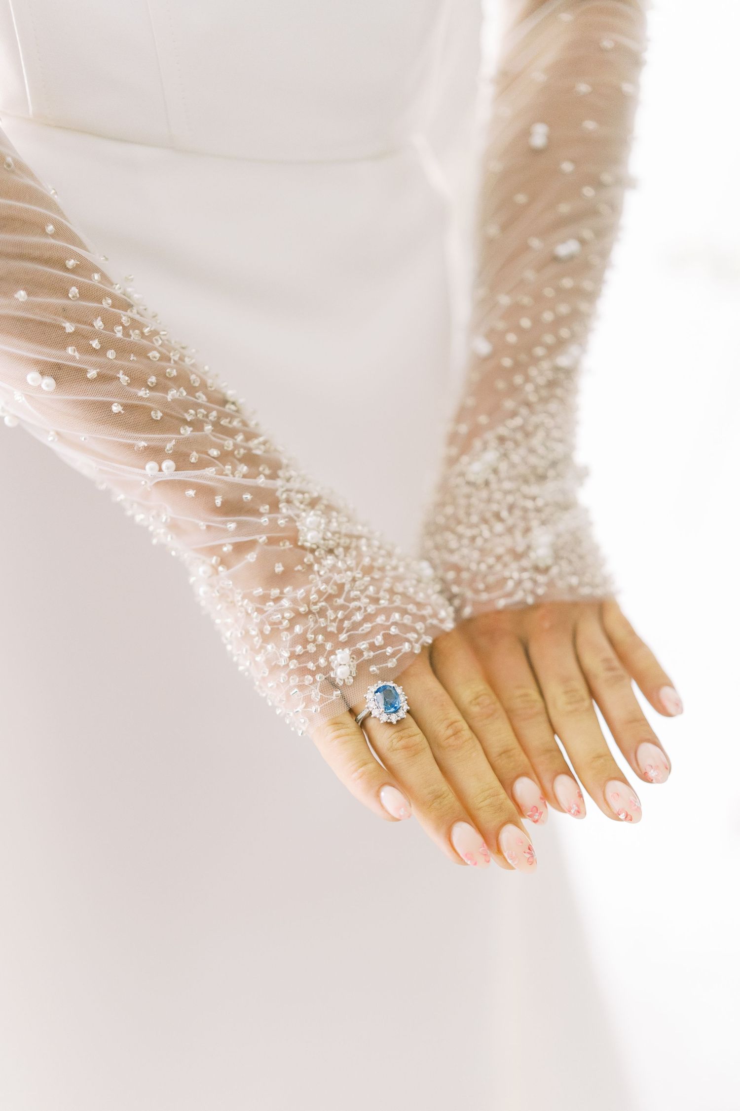 Close-up of bride's hands wearing pearl-embellished gloves and a blue sapphire engagement ring.
