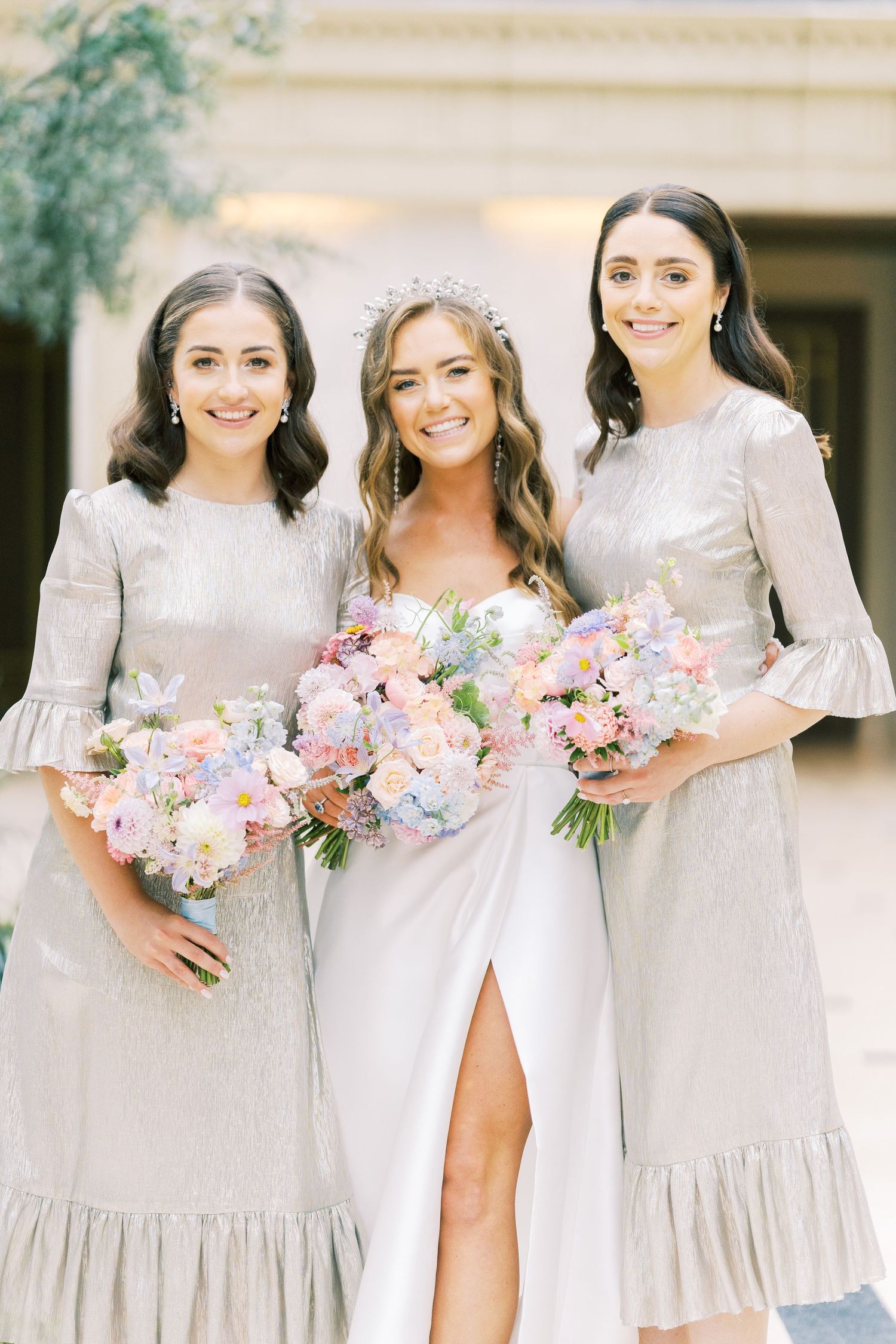 Bride in white gown with tiara flanked by two bridesmaids in silver dresses holding pastel floral bouquets.