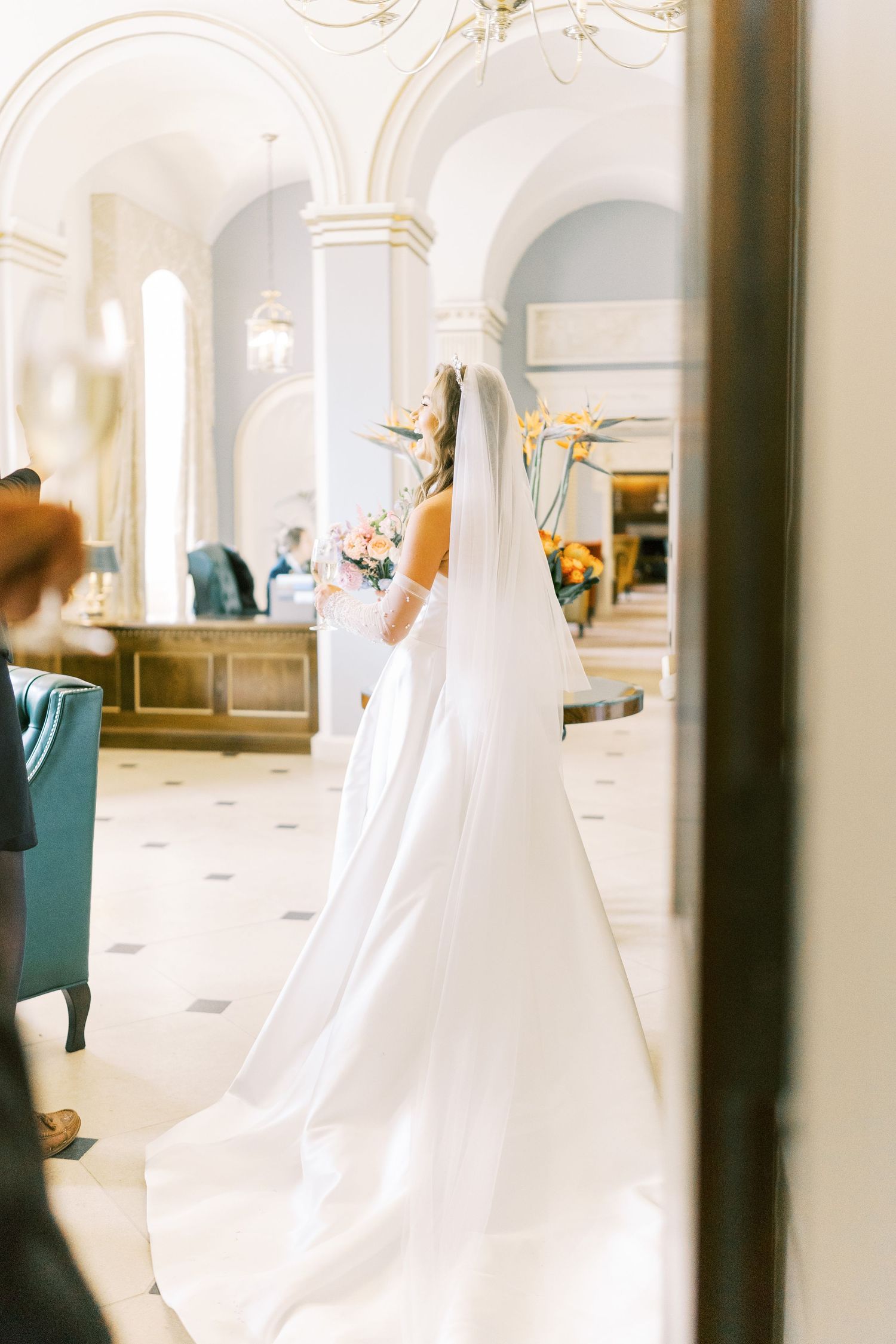 Bride in white gown and veil holding bouquet stands in elegant venue with arched ceilings and marble floors.