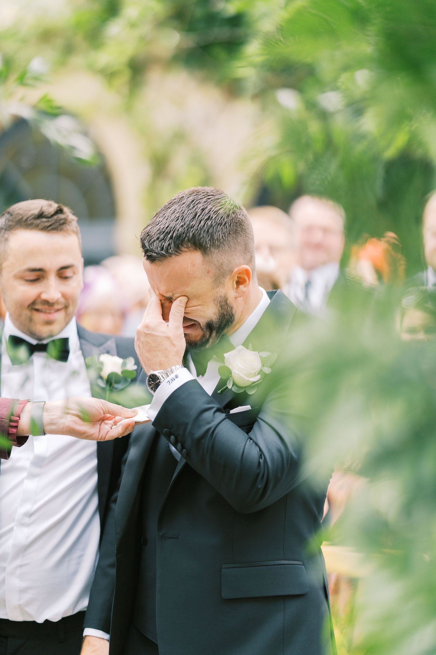 Emotional groom wiping tears at wedding ceremony, wearing dark suit with white boutonniere, surrounded by groomsmen.