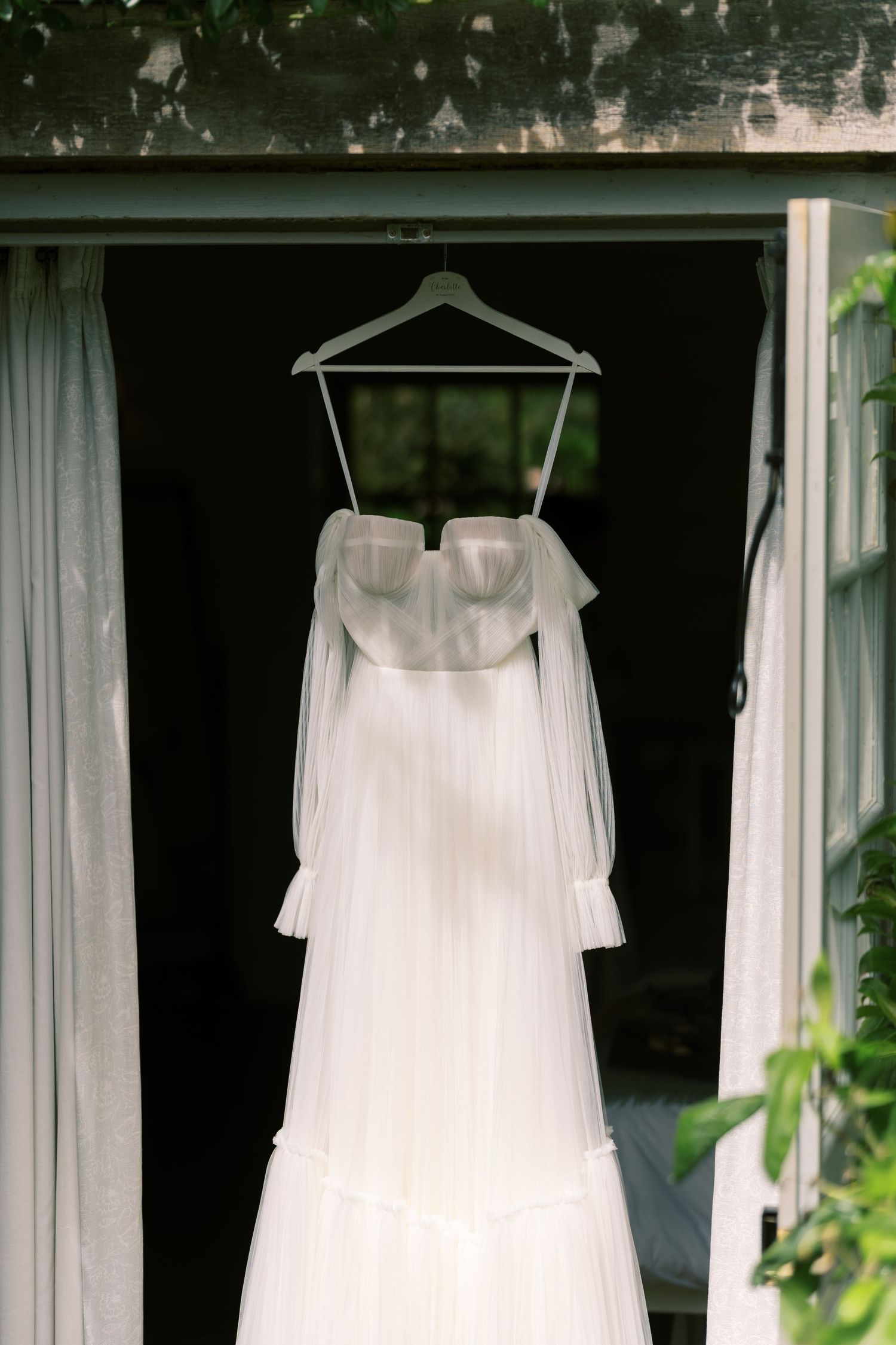 White off-shoulder wedding dress with long sleeves hanging on a hanger in a rustic doorway.
