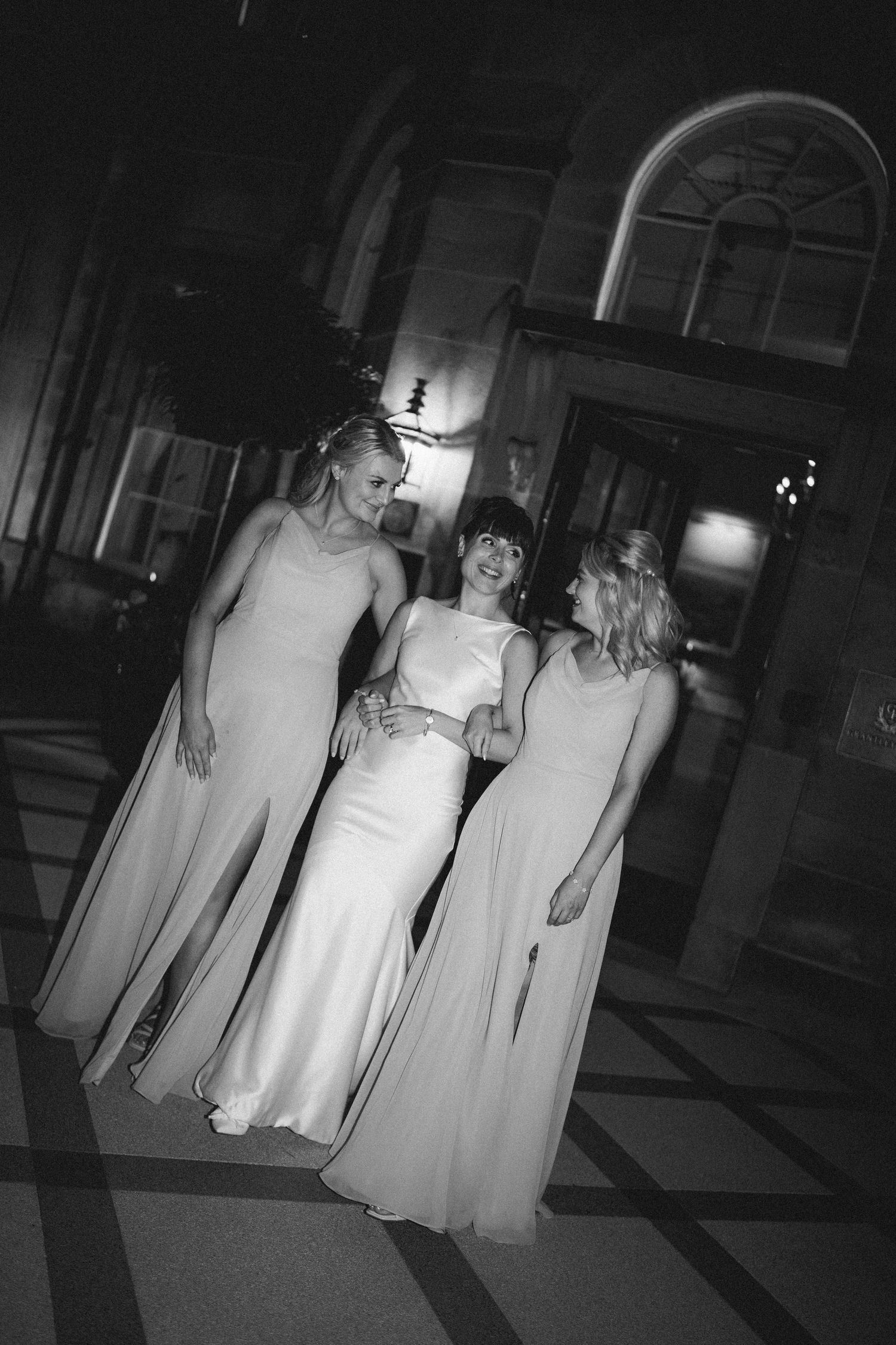 Black and white photo of bride and two bridesmaids in elegant gowns outside a grand venue.