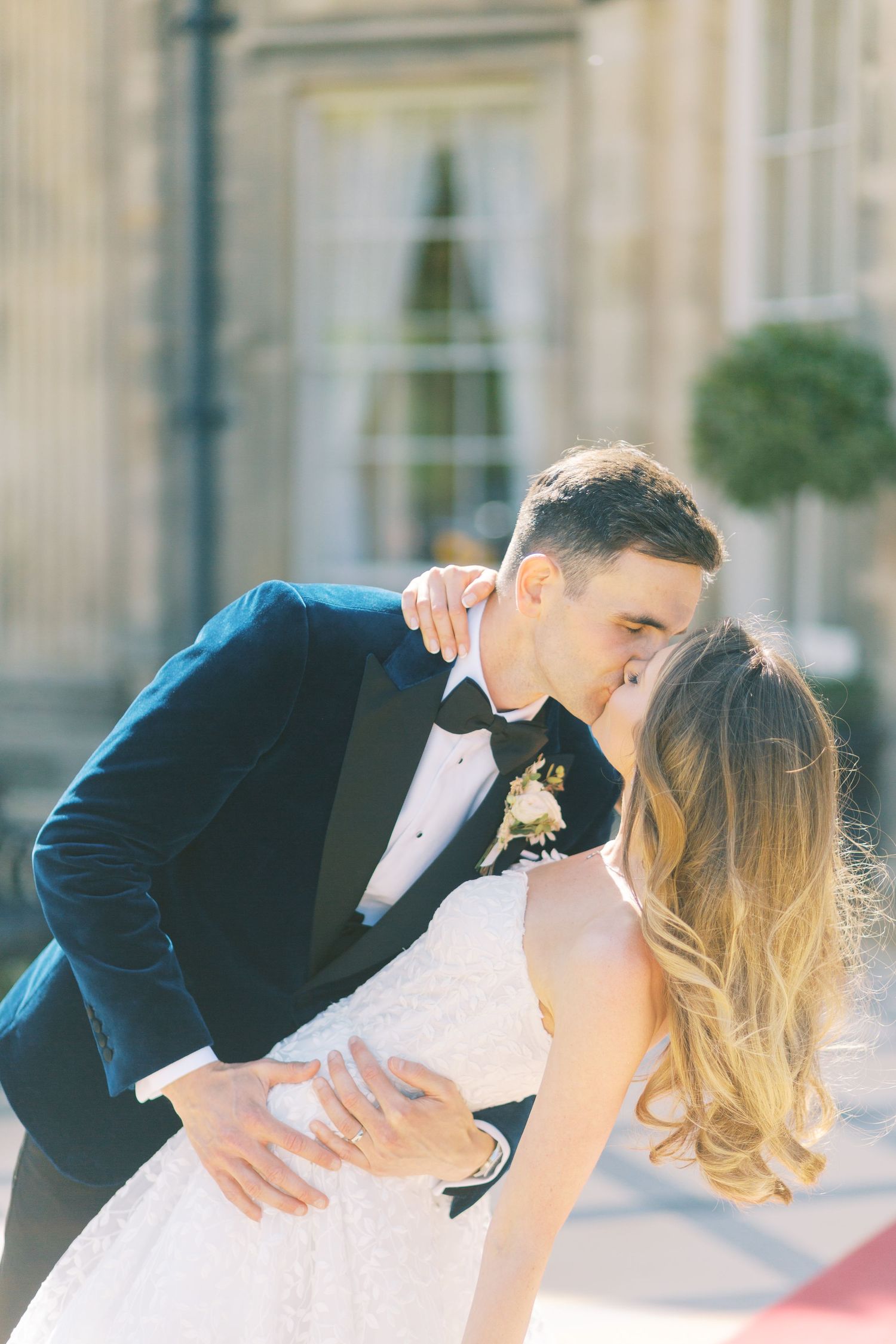 A bride and groom share a romantic dip kiss outdoors, dressed in a white wedding gown and navy tuxedo.