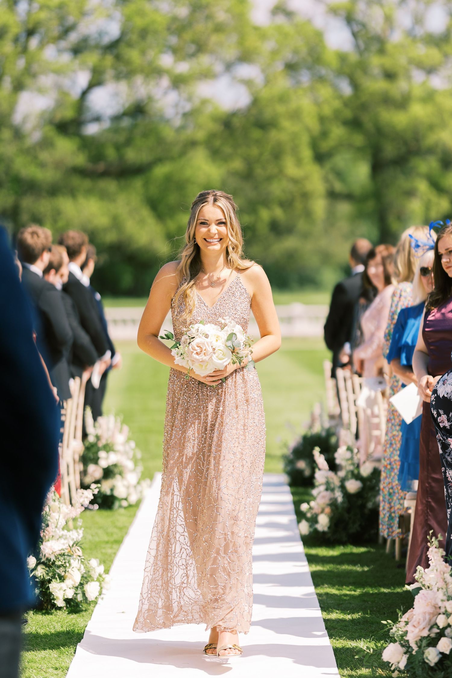 Smiling bridesmaid walks down outdoor wedding aisle in rose gold sequin gown holding white floral bouquet.