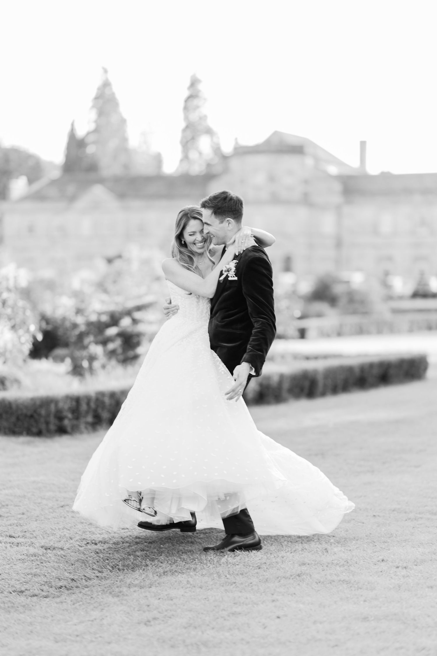 Black and white photo of bride and groom dancing joyfully in garden with grand estate building backdrop.