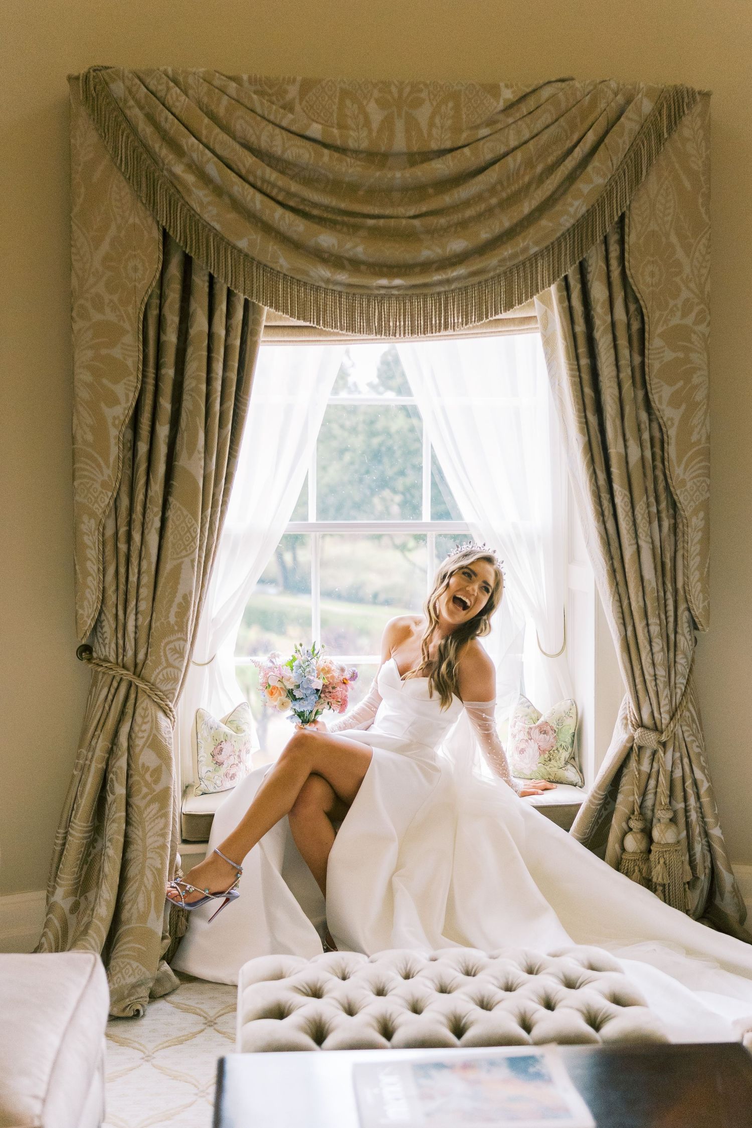 Bride in white gown sits on tufted window seat holding colorful bouquet, laughing joyfully in elegant draped room.