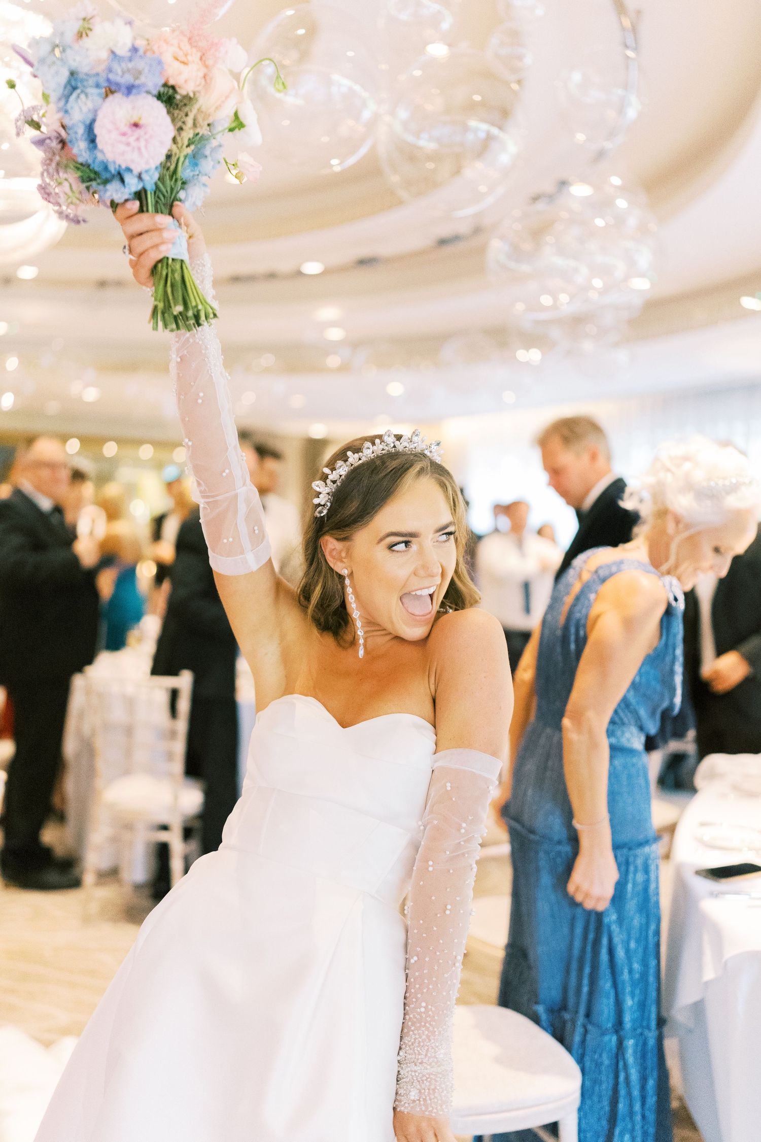 Joyful bride in strapless gown and tiara raises floral bouquet while celebrating at elegant wedding reception.