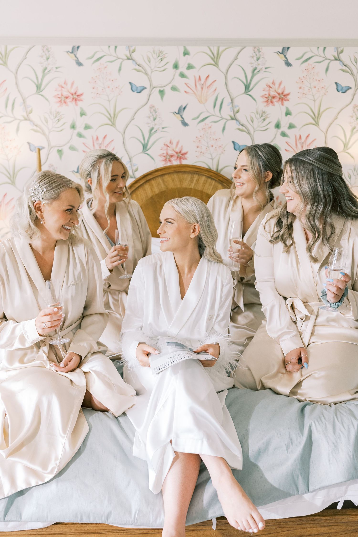 Bride and bridesmaids laughing together on bed in matching robes before wedding ceremony.