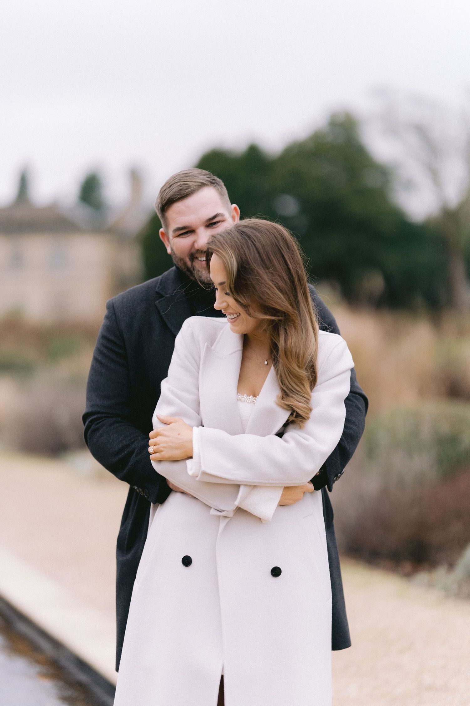 Joyful couple embraces outdoors, man hugging woman from behind near a tranquil garden pathway.