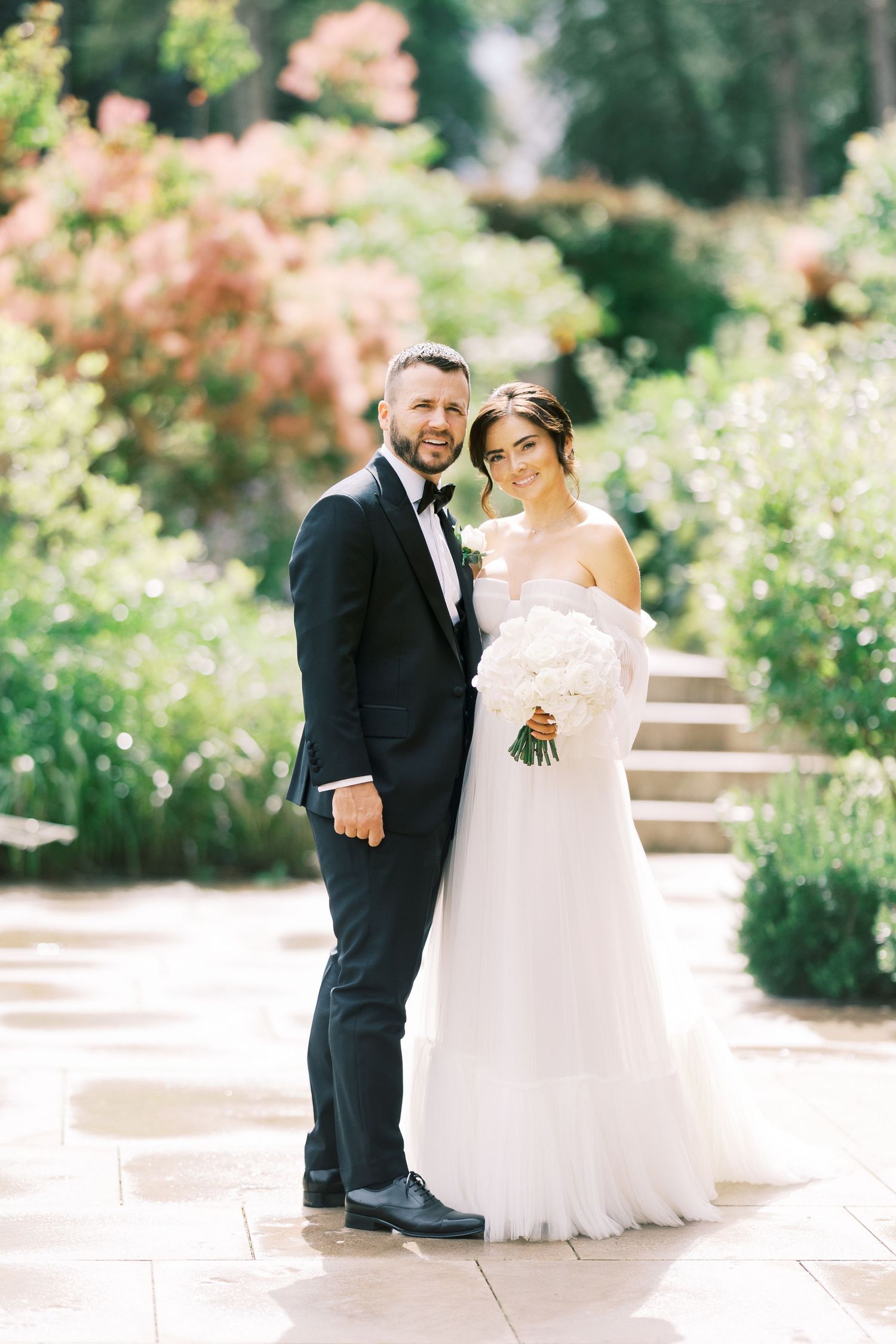 Bride and groom posing in lush garden, bride in strapless tulle gown holding white bouquet, groom in black tuxedo.