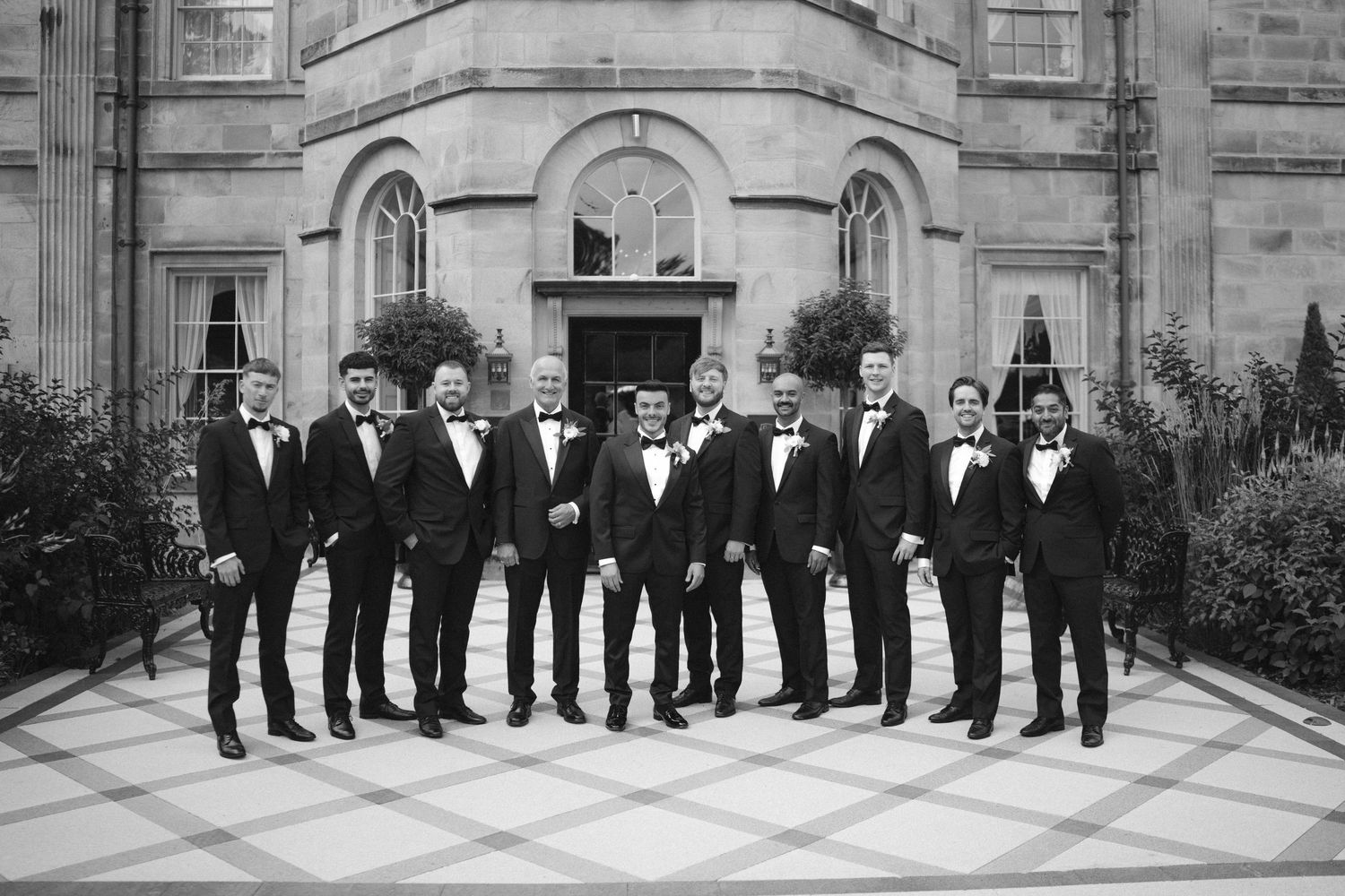 Black and white photo of groom and groomsmen in tuxedos posing outside an elegant stone building.