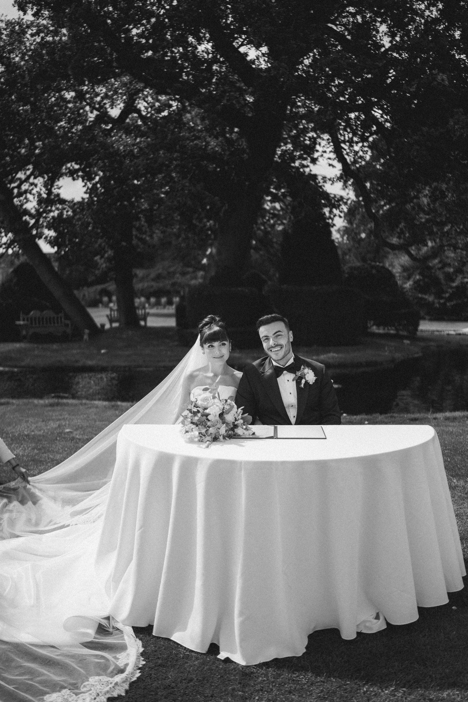Bride and groom smiling at signing table during outdoor wedding ceremony in black and white.