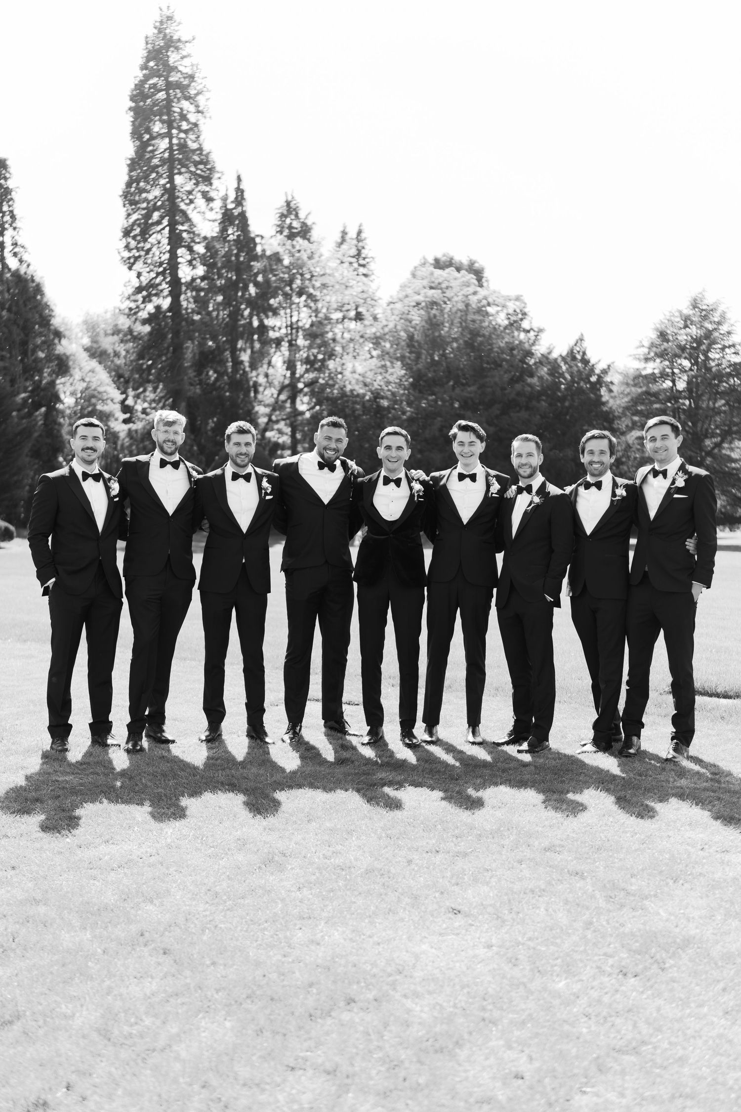 A groom and his groomsmen pose in matching black tuxedos and bow ties outdoors in a black and white photo.