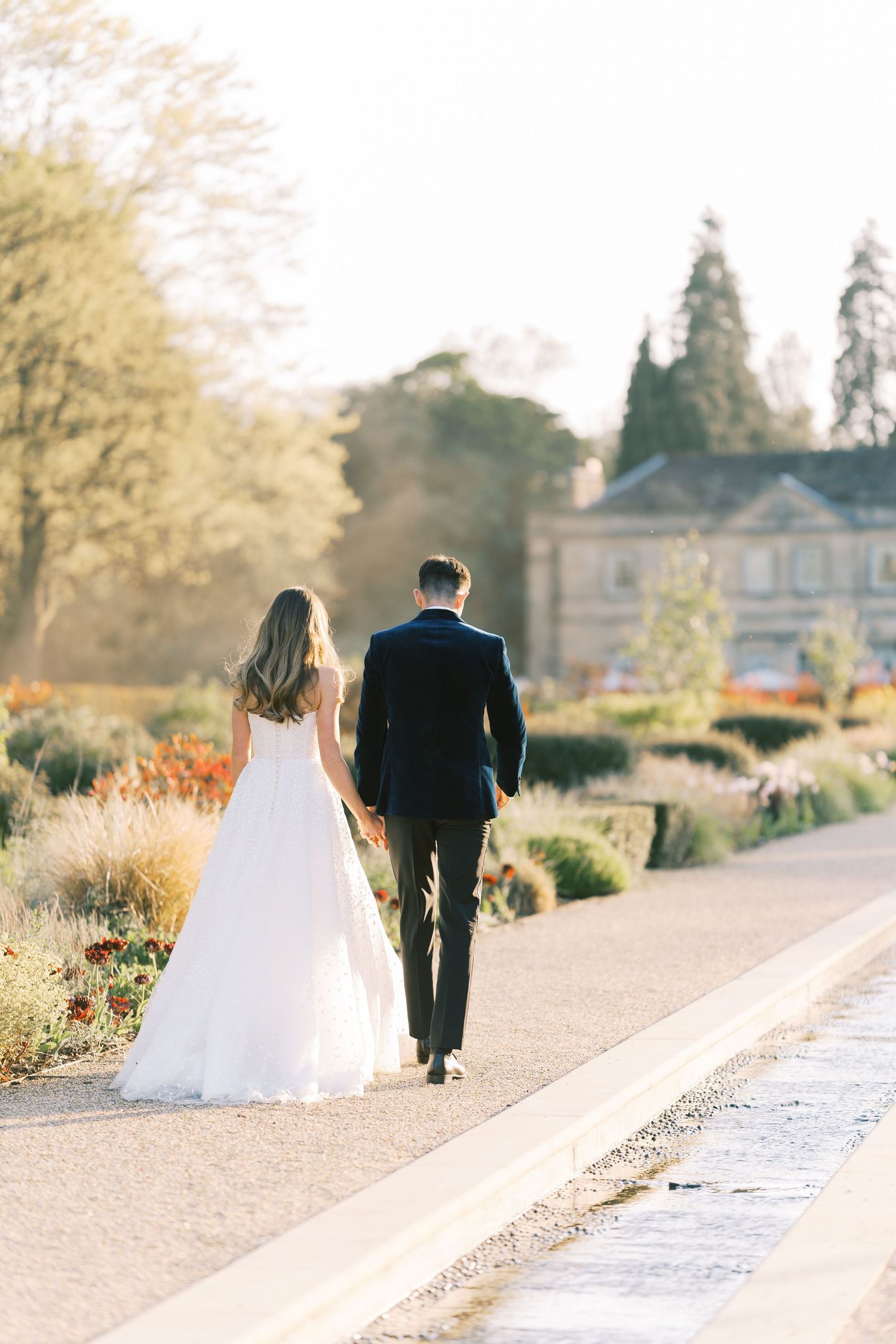 A bride and groom walk hand in hand along a sunlit garden path toward an elegant estate on their wedding day.