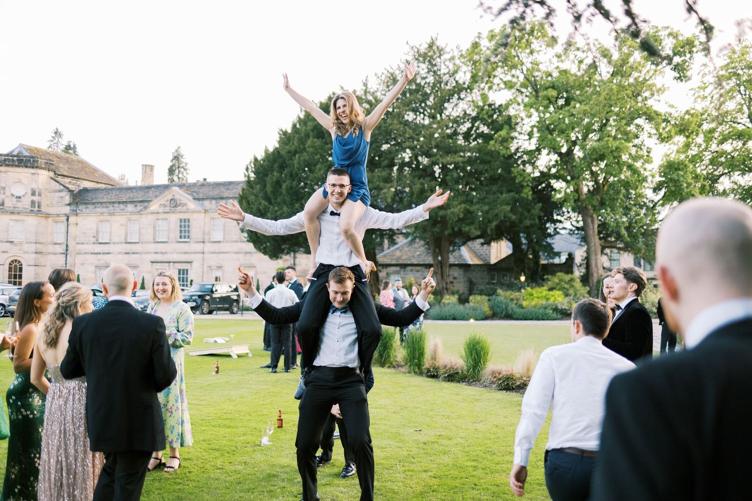 A woman lifted on shoulders celebrates outdoors at an elegant wedding reception on a manicured lawn.