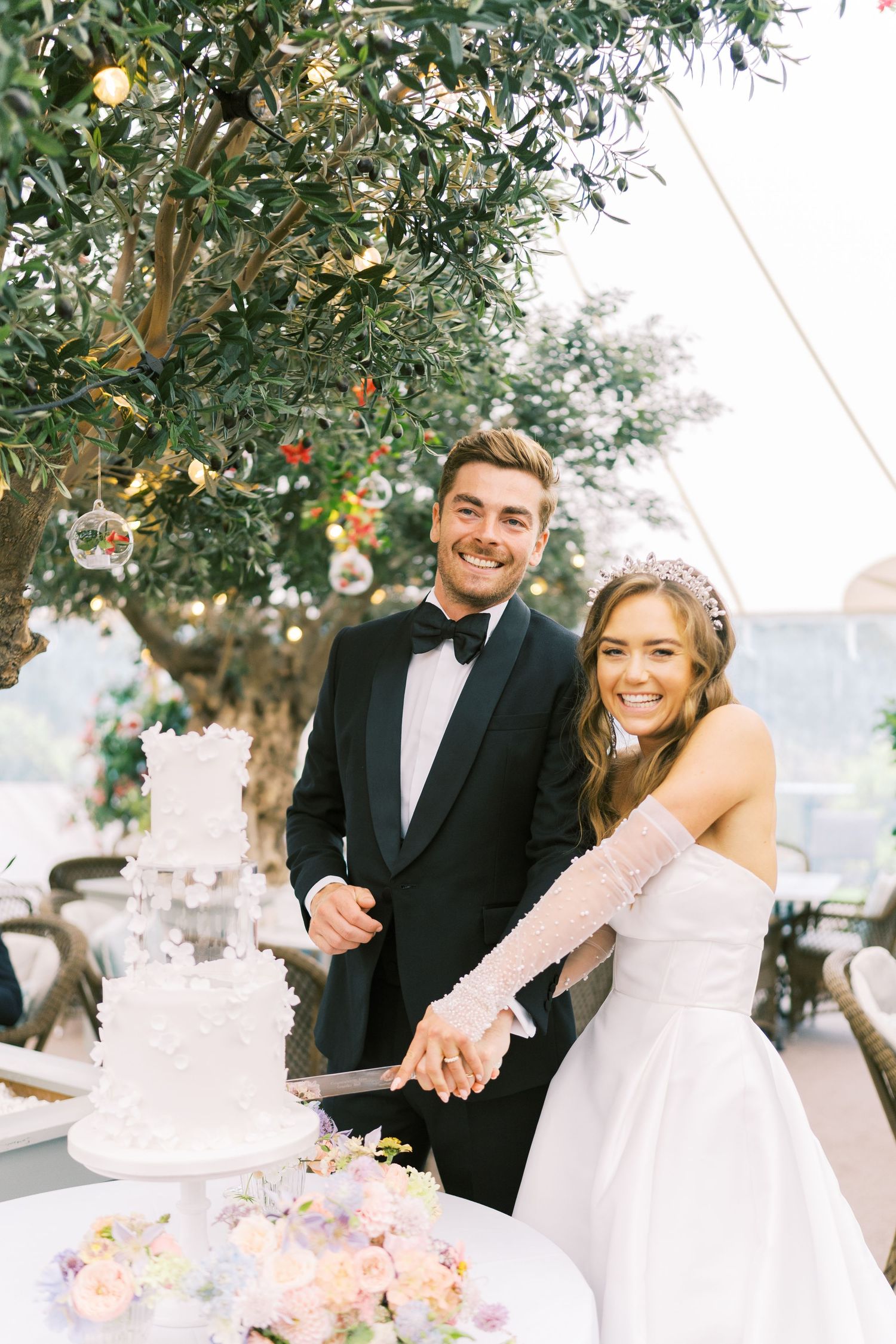Smiling bride and groom cutting white tiered wedding cake outdoors under olive tree with string lights.