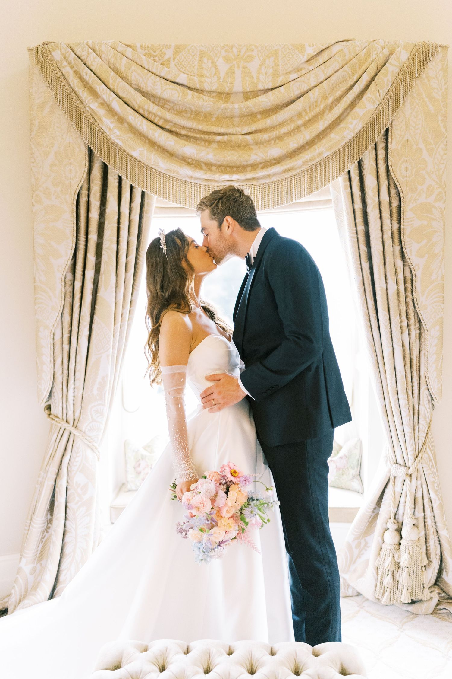 Bride and groom share romantic kiss in front of ornate draped window, bride holding pastel floral bouquet.
