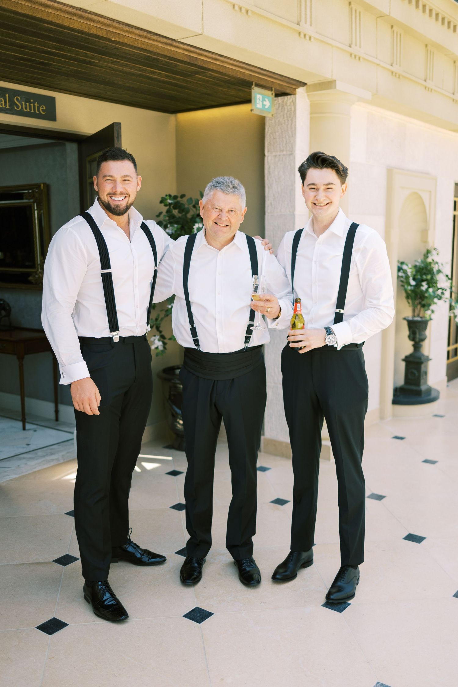 Three men in white shirts and black suspenders smiling together outside a hotel suite.