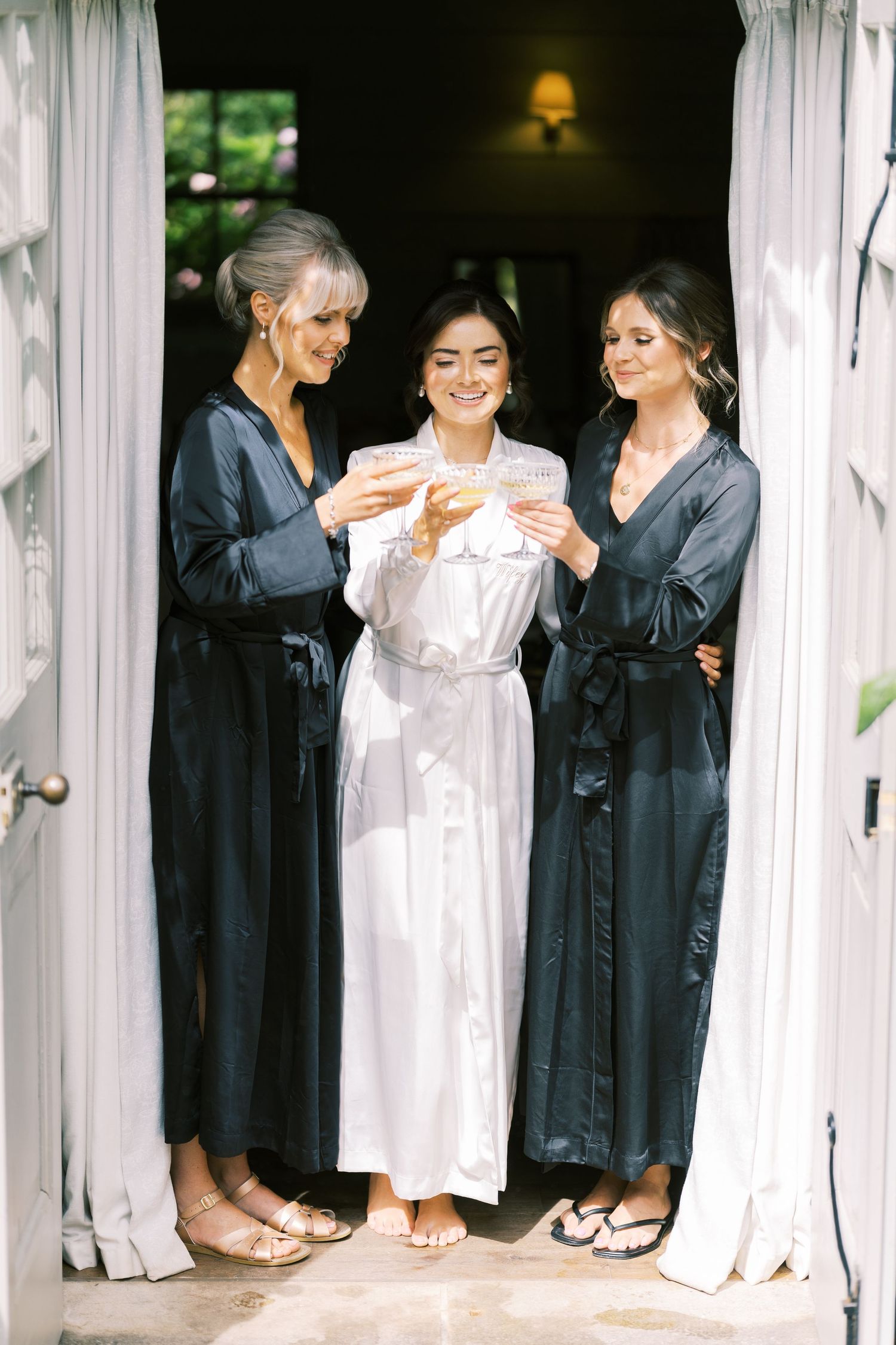 Bride in white robe celebrates with two bridesmaids in black satin robes in a doorway.