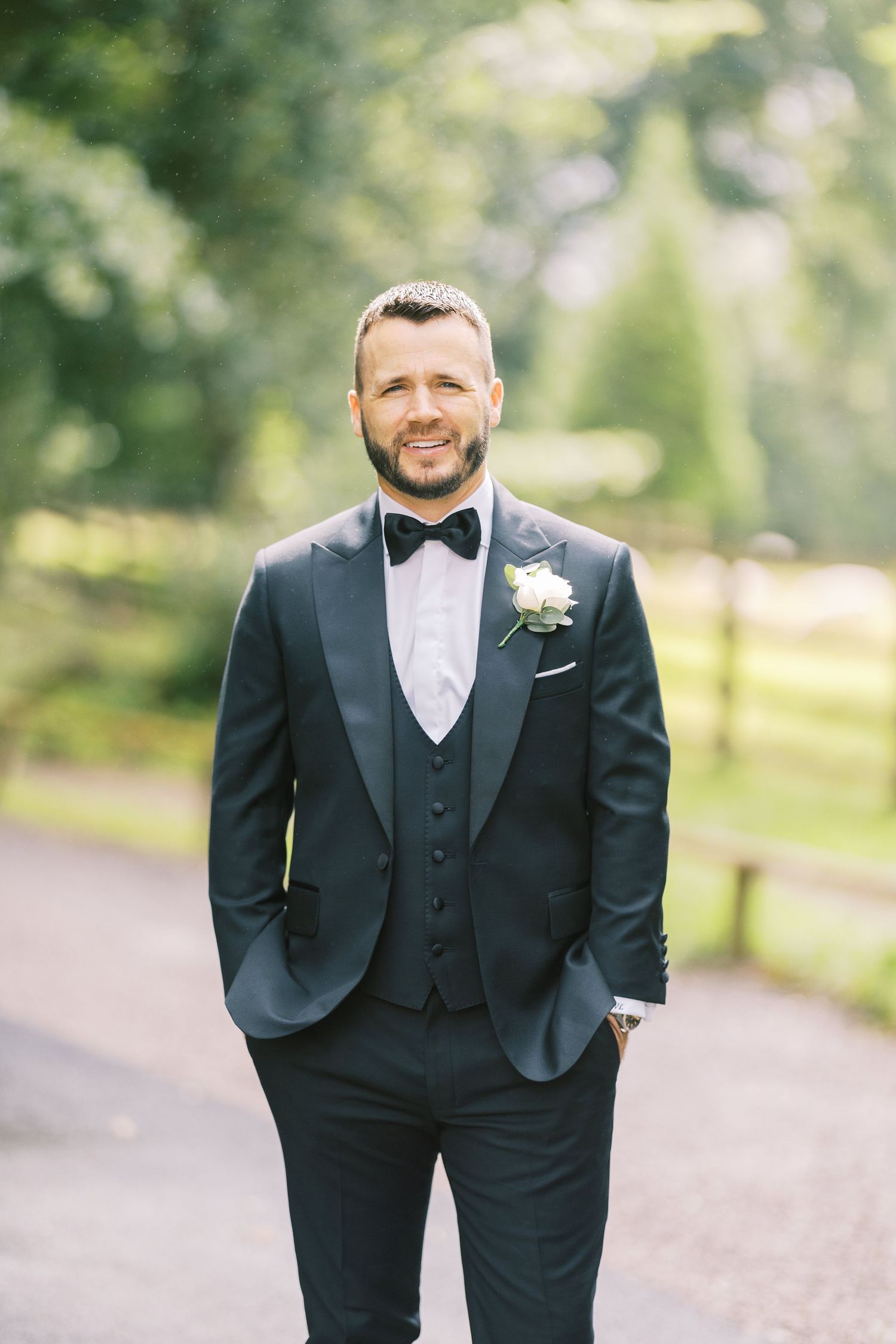 Smiling groom in dark navy tuxedo with bow tie and white boutonniere stands outdoors on a tree-lined path.