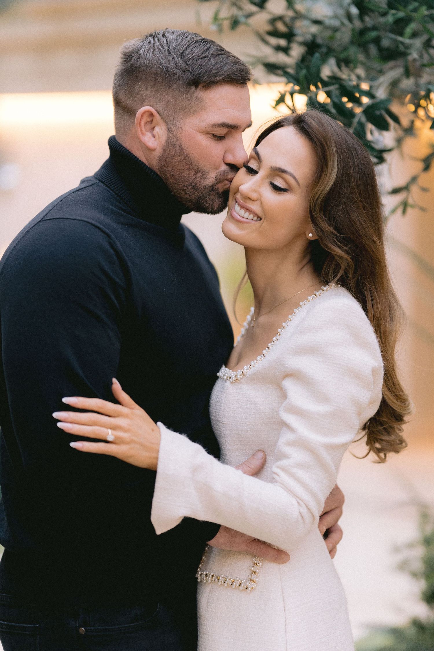 Man in black turtleneck kissing smiling woman in white tweed dress during romantic outdoor photo session.