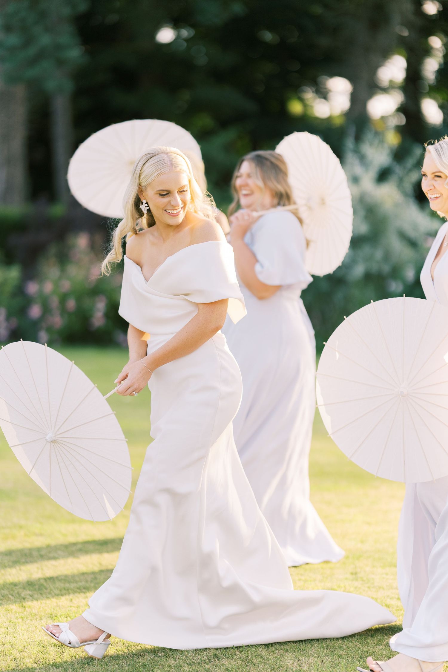 Bride and bridesmaids in white gowns twirling parasols on sunlit garden lawn.