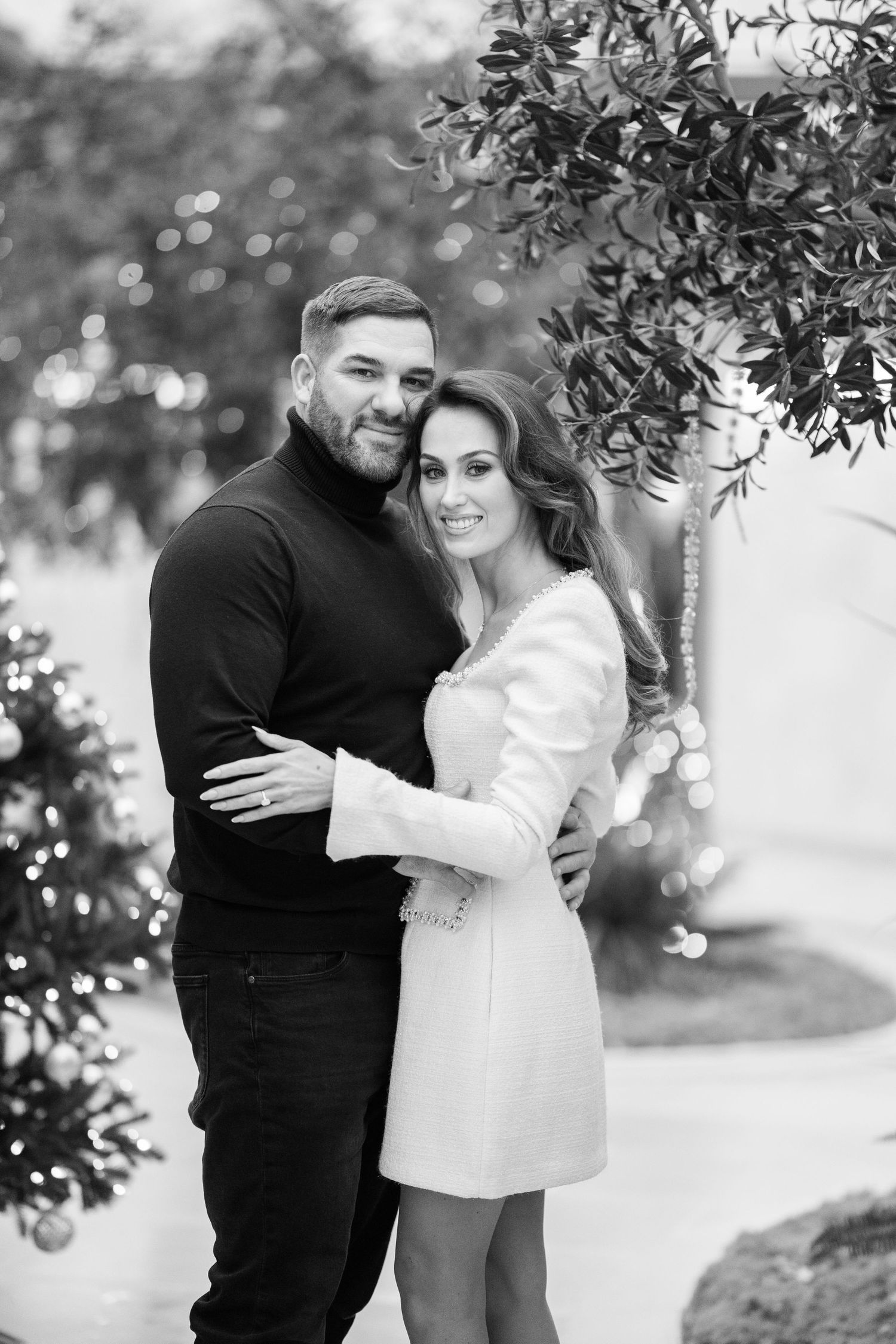 Black and white portrait of a loving couple embracing outdoors near festive Christmas trees with bokeh lights.