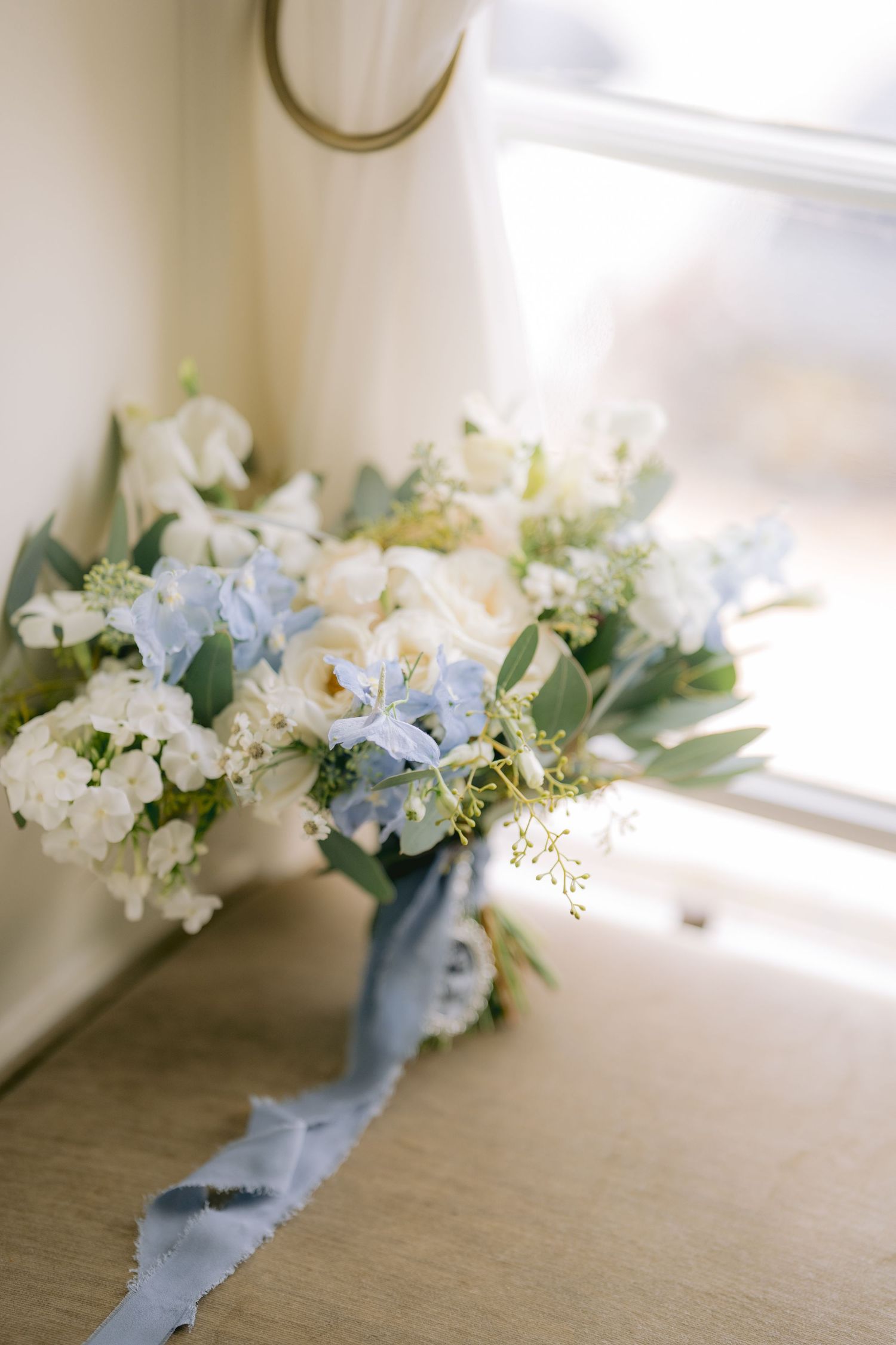 Elegant wedding bouquet with white roses and blue flowers tied with dusty blue silk ribbon near a sunlit window.