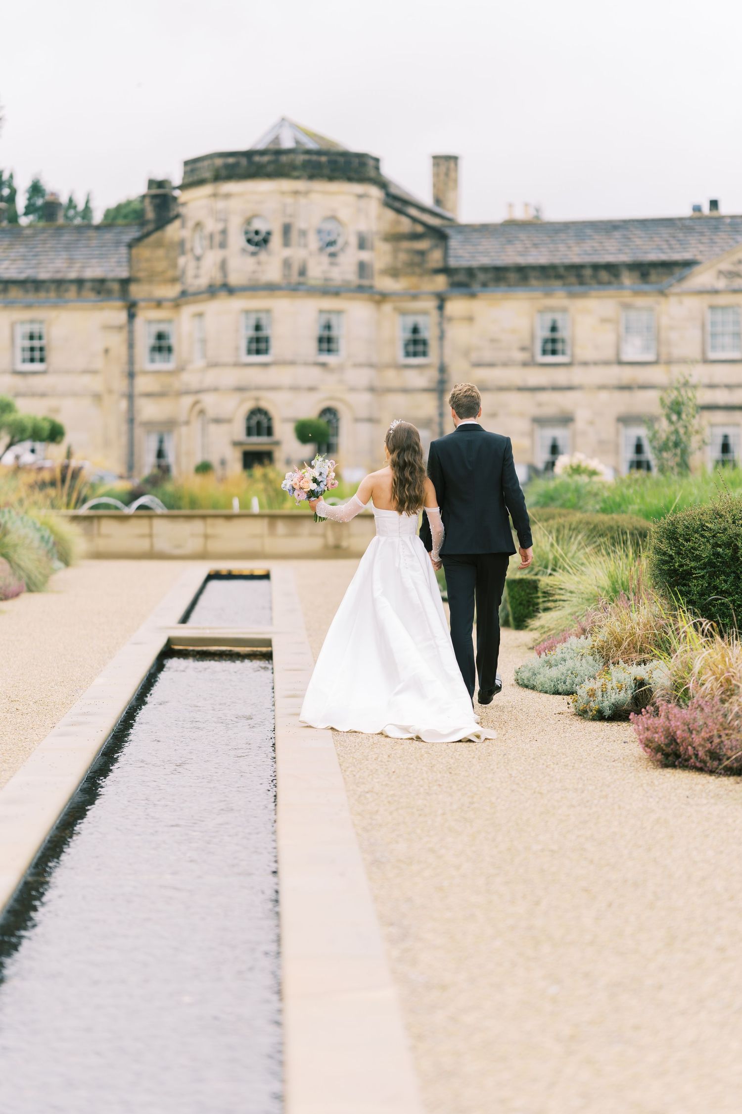 Bride and groom walk hand in hand toward a grand stone manor along a garden path with a reflecting pool.