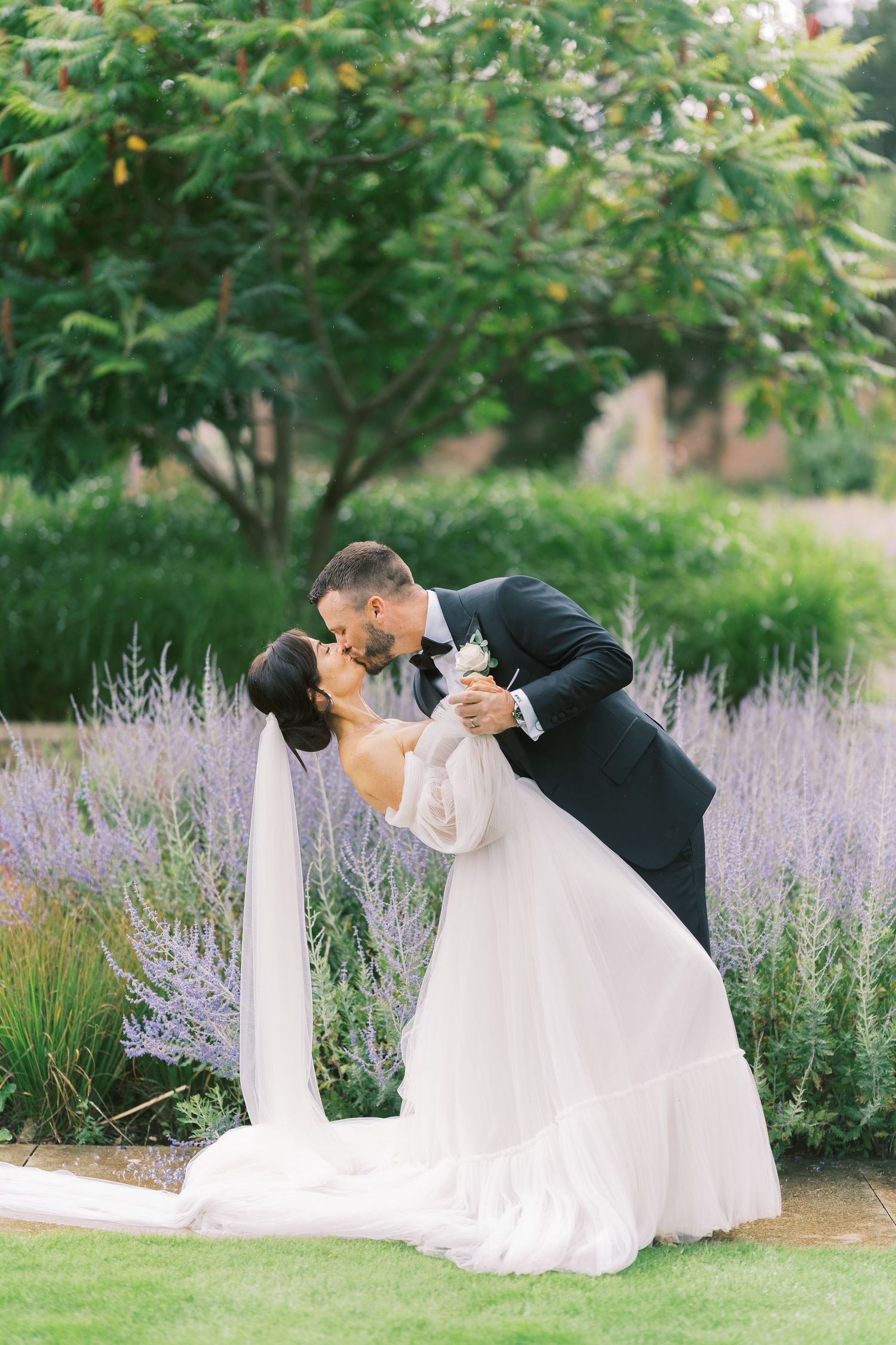 Bride and groom share a romantic dip kiss surrounded by lavender blooms and lush greenery in an elegant garden setting.