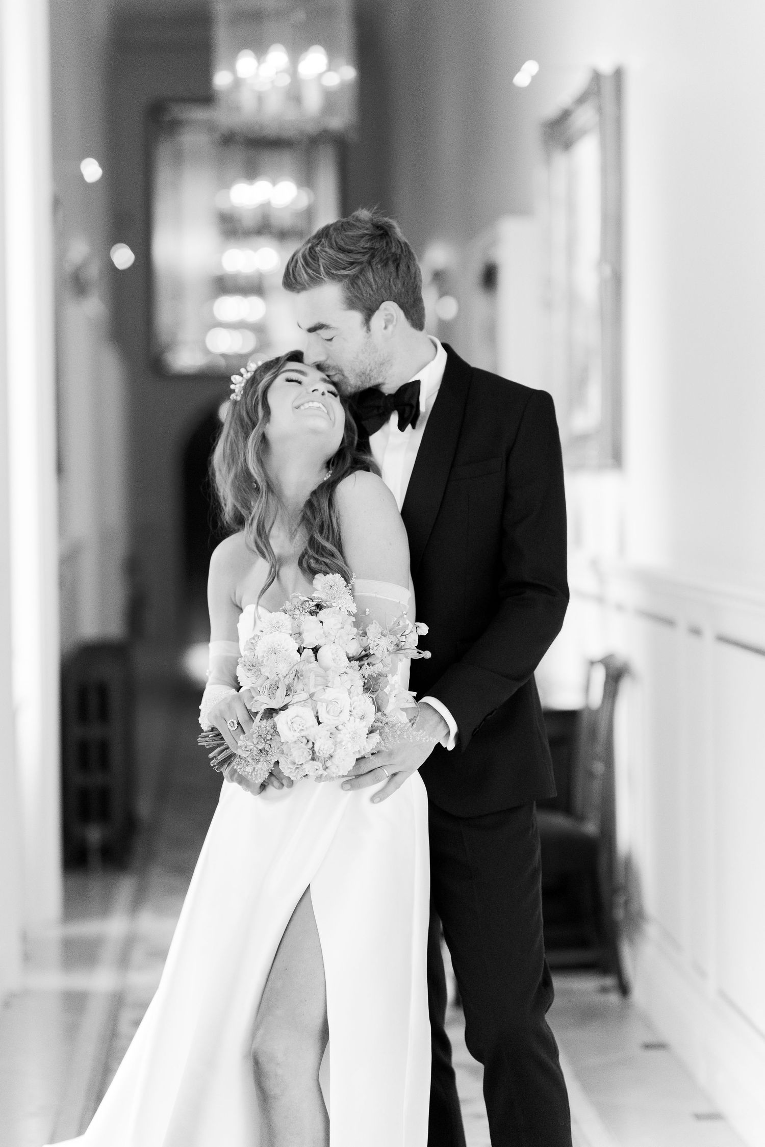 Black and white wedding photo of bride and groom embracing in elegant hallway, bride holding floral bouquet.
