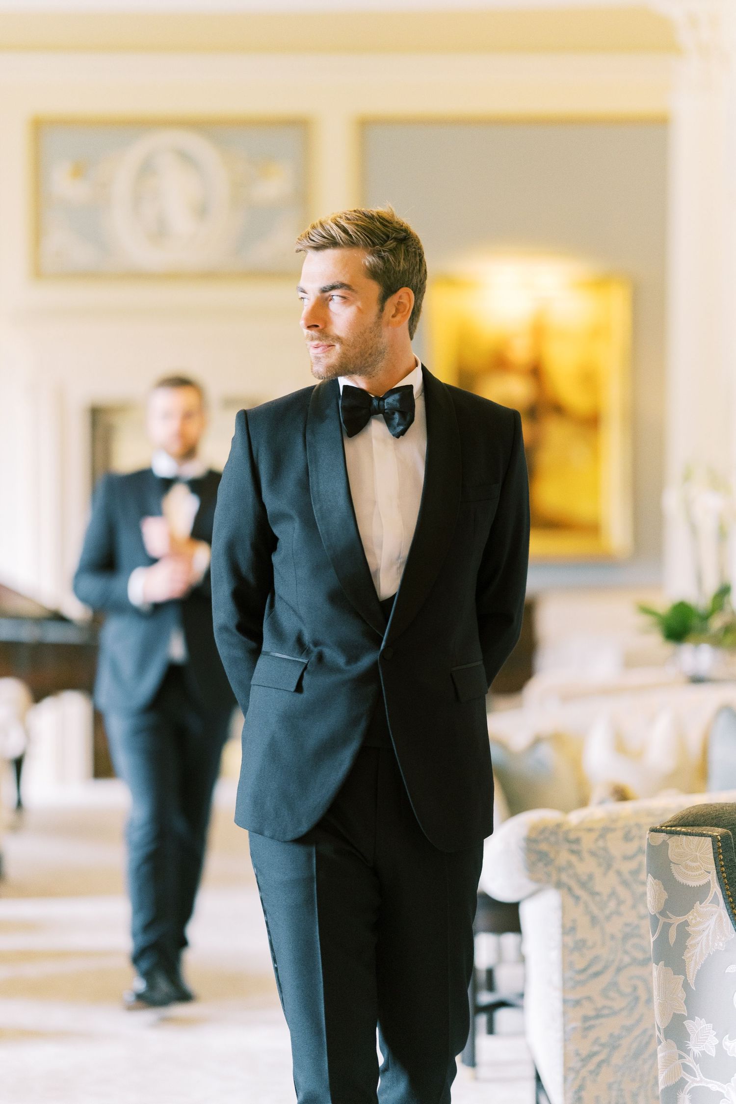 Elegant groom in black tuxedo with bow tie standing confidently in a luxurious wedding venue.