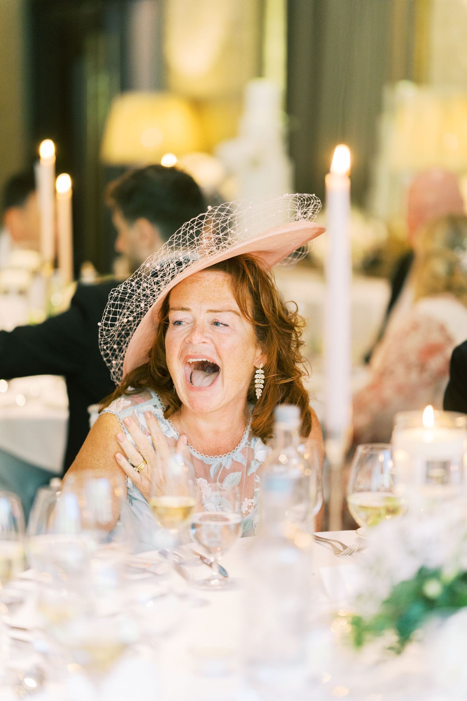 Laughing female wedding guest wearing pink fascinator hat at elegant candlelit reception dinner.