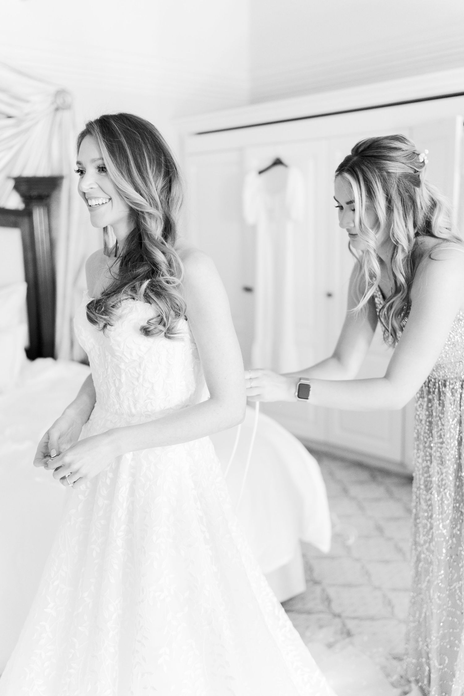 Black and white photo of a bride smiling in a lace wedding gown while a bridesmaid helps fasten her dress.