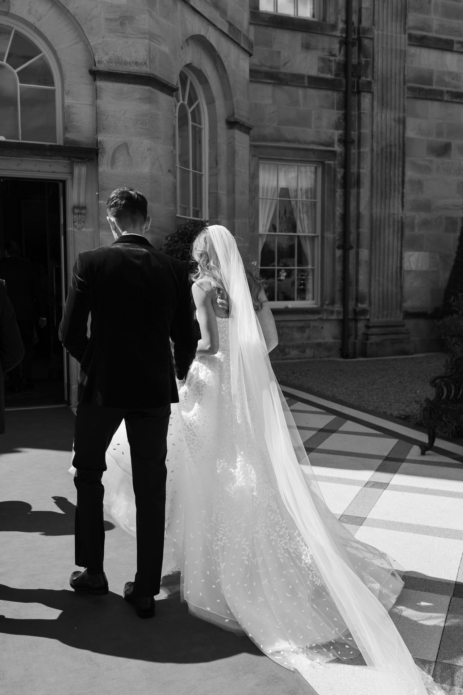 Bride and groom walk hand in hand outside stone mansion in black and white photo.