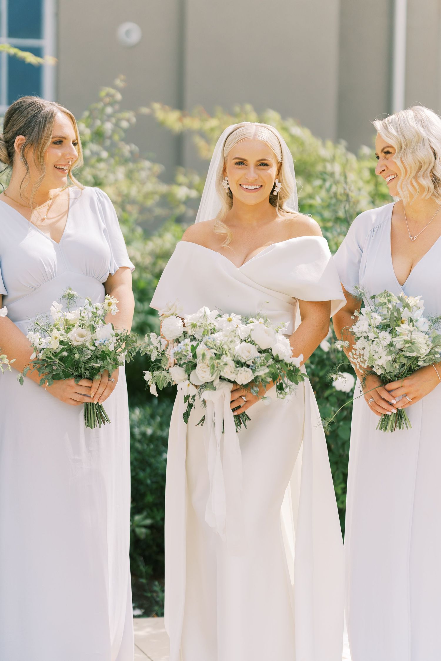Bride in off-shoulder white gown with veil holds bouquet, flanked by two bridesmaids in light blue dresses with white flowers.