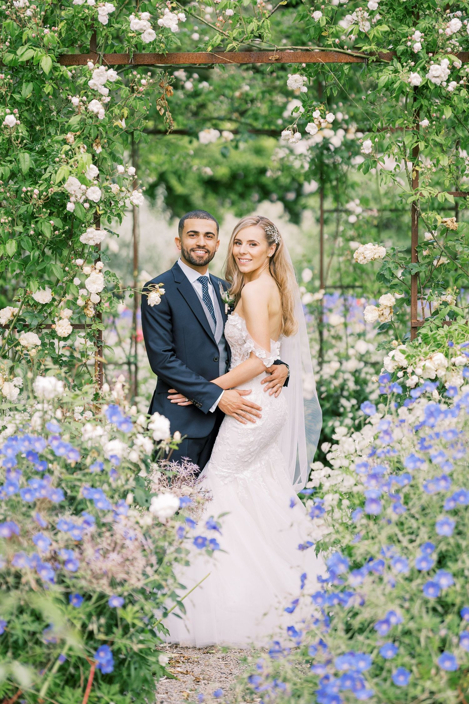 A bride and groom embrace under a floral arch surrounded by blue and white flowers in a lush garden setting.