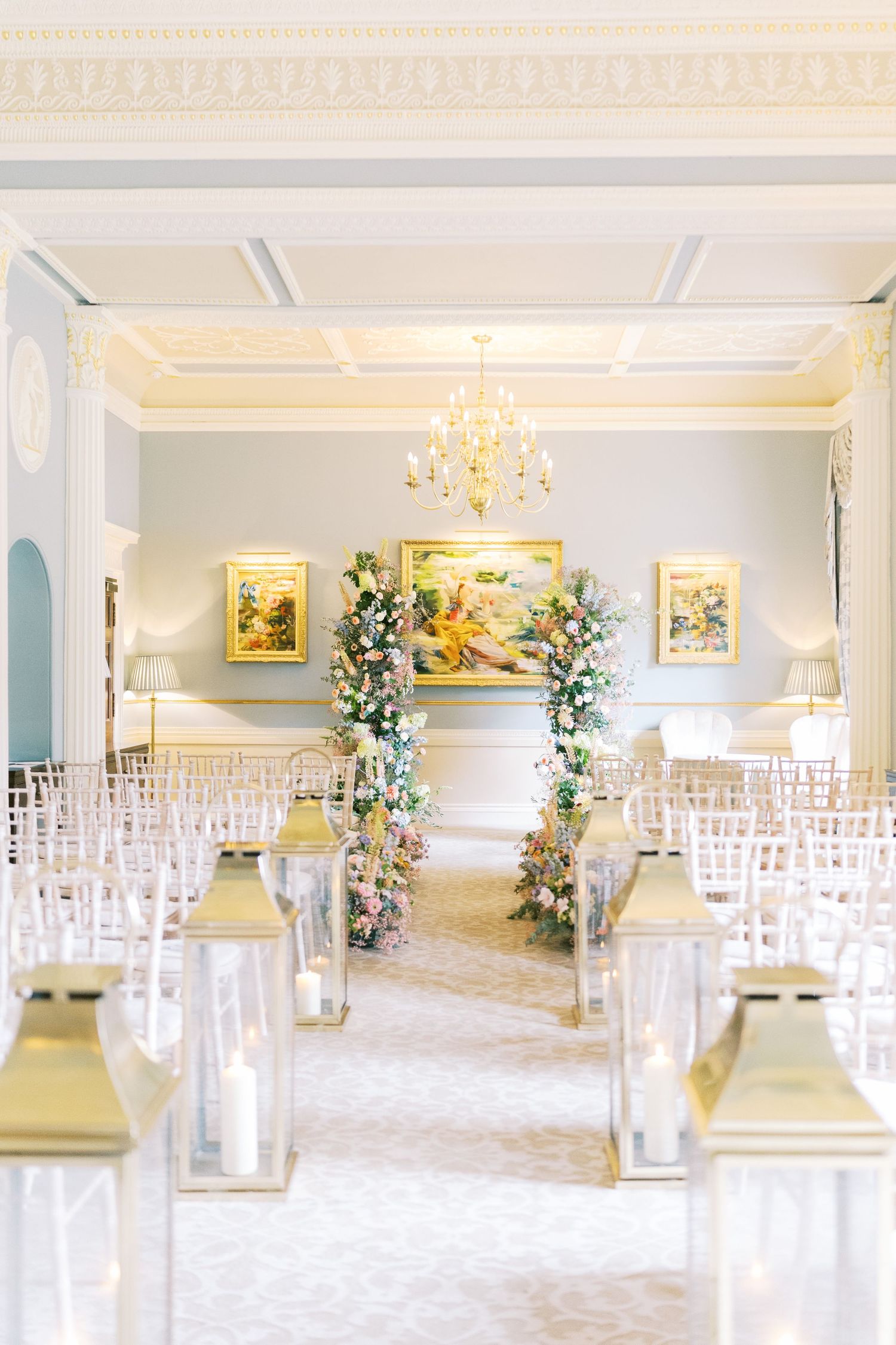 Elegant wedding ceremony room with ghost chairs, gold lanterns, floral arch, chandelier, and soft blue walls.