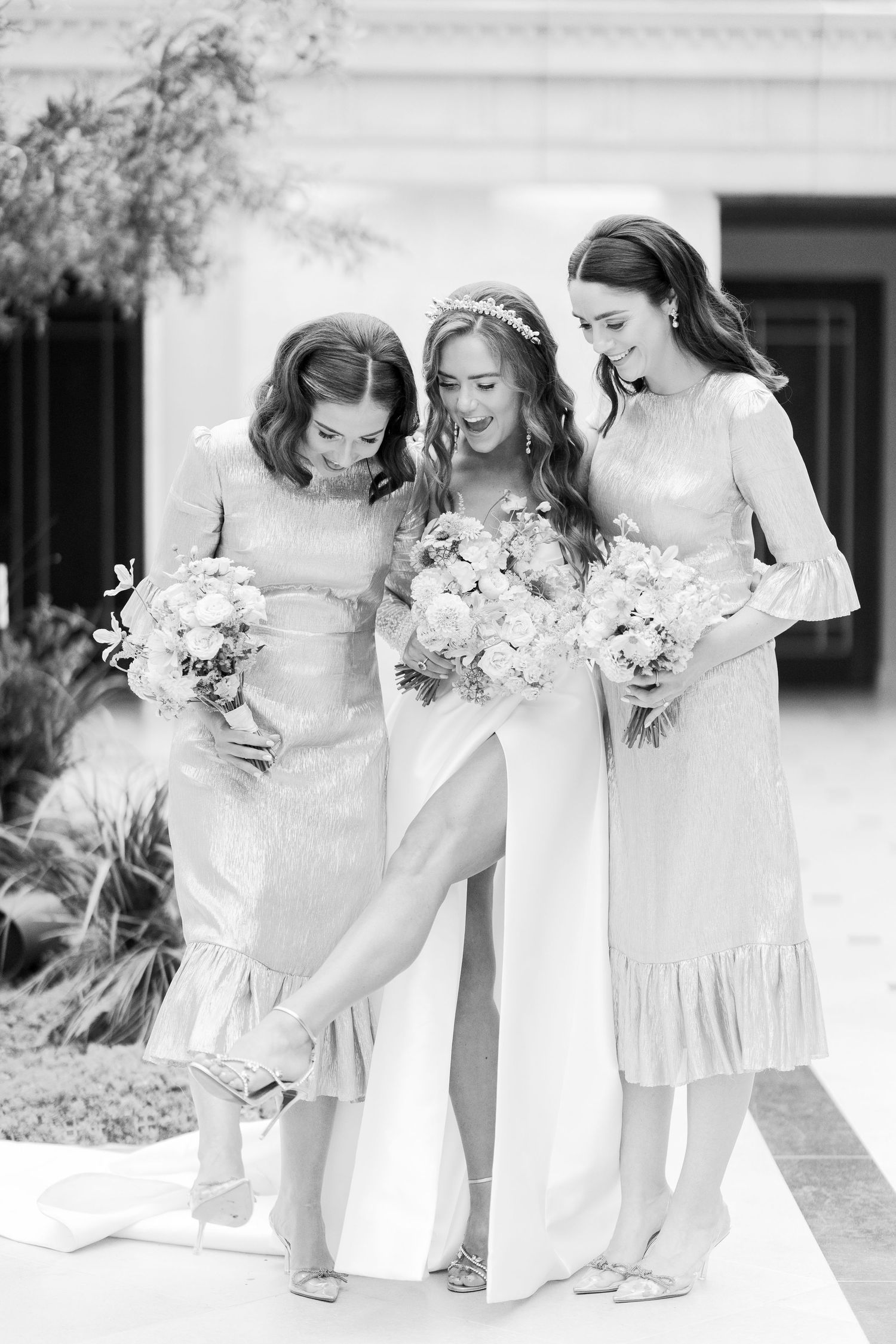 Black and white photo of a bride and two bridesmaids laughing together while holding bouquets outdoors.